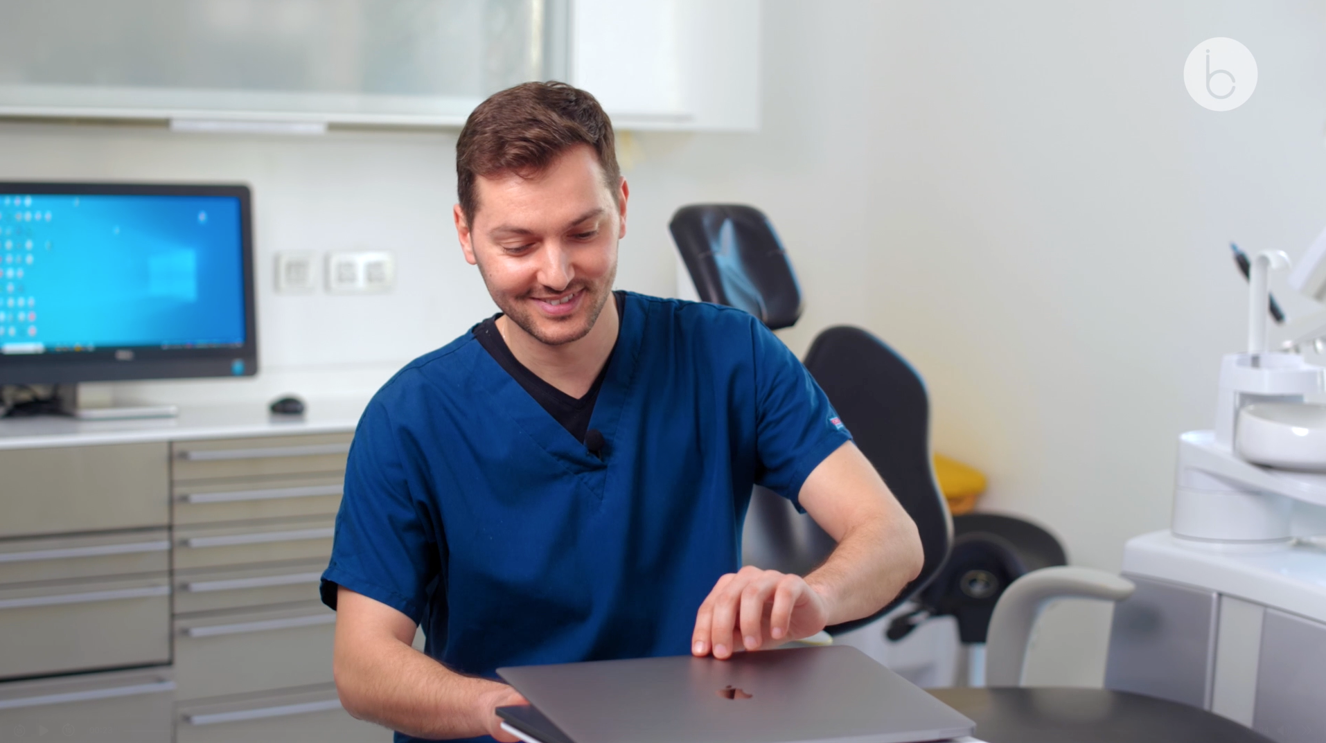 A male healthcare professional in blue scrubs smiling and working on a laptop in a medical office.