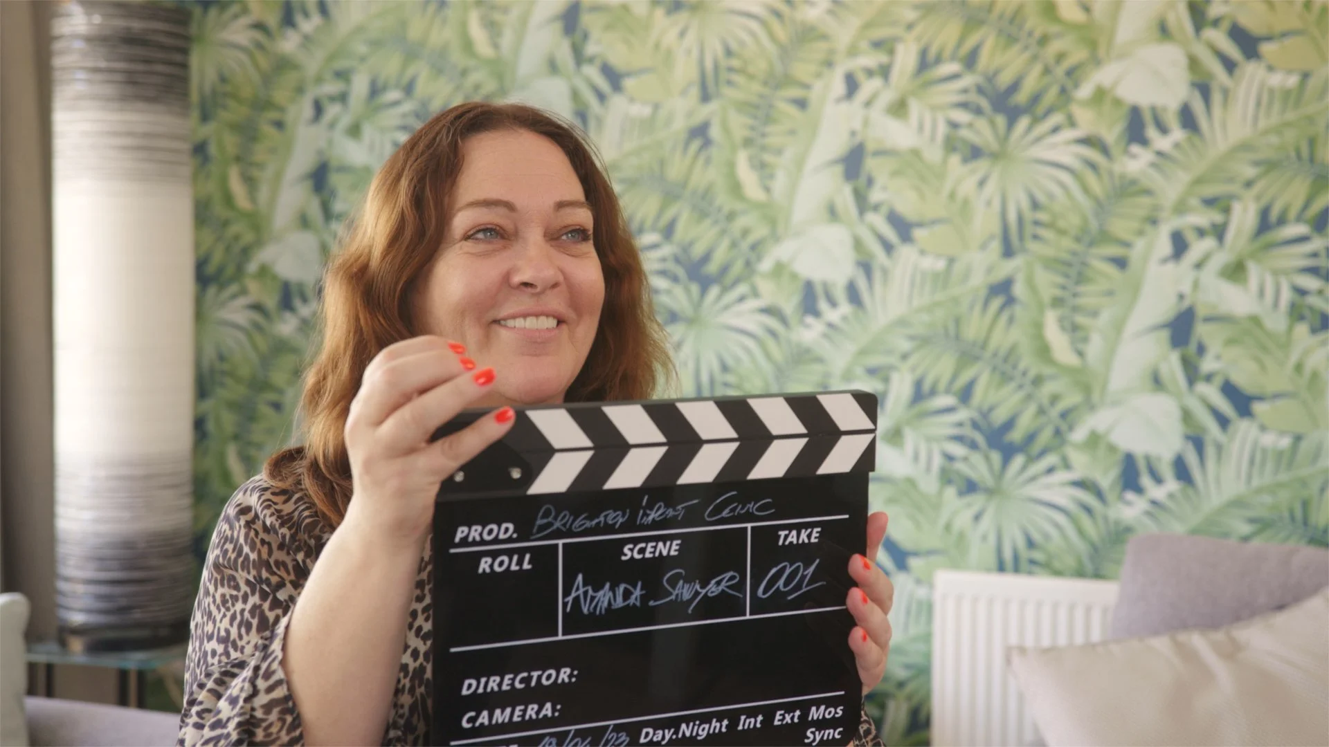 A woman with red hair and a leopard print top holding a film slate in a room with green botanical wallpaper in the background.