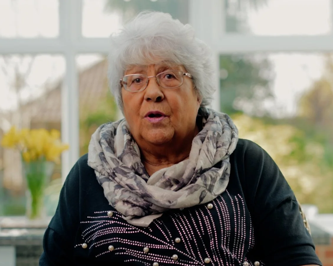 An elderly woman with white hair and glasses sitting inside, wearing a black top with white dots and pearls and a gray patterned scarf, with a blurred background of windows and greenery.