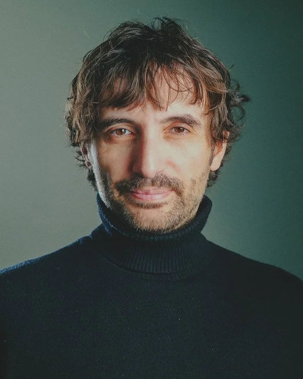 A man with shaggy brown hair and a beard wearing a black turtleneck sweater, looking at the camera with a slight smile, against a plain, muted background.