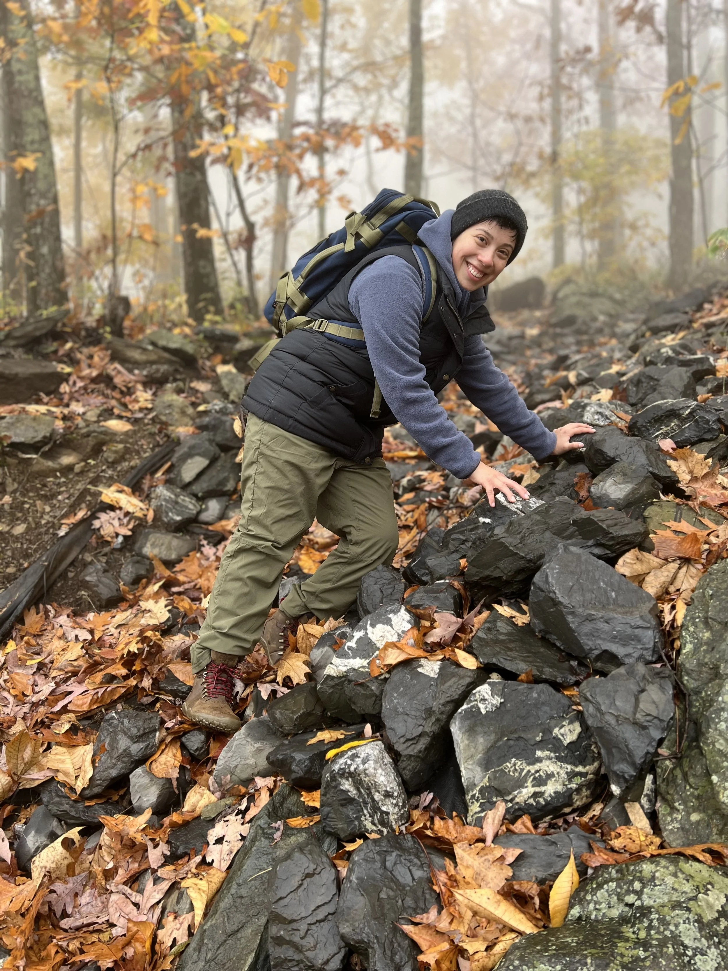 Person climbing over rocks on a forest trail in autumn, wearing a beanie, backpack, and outdoor clothing.