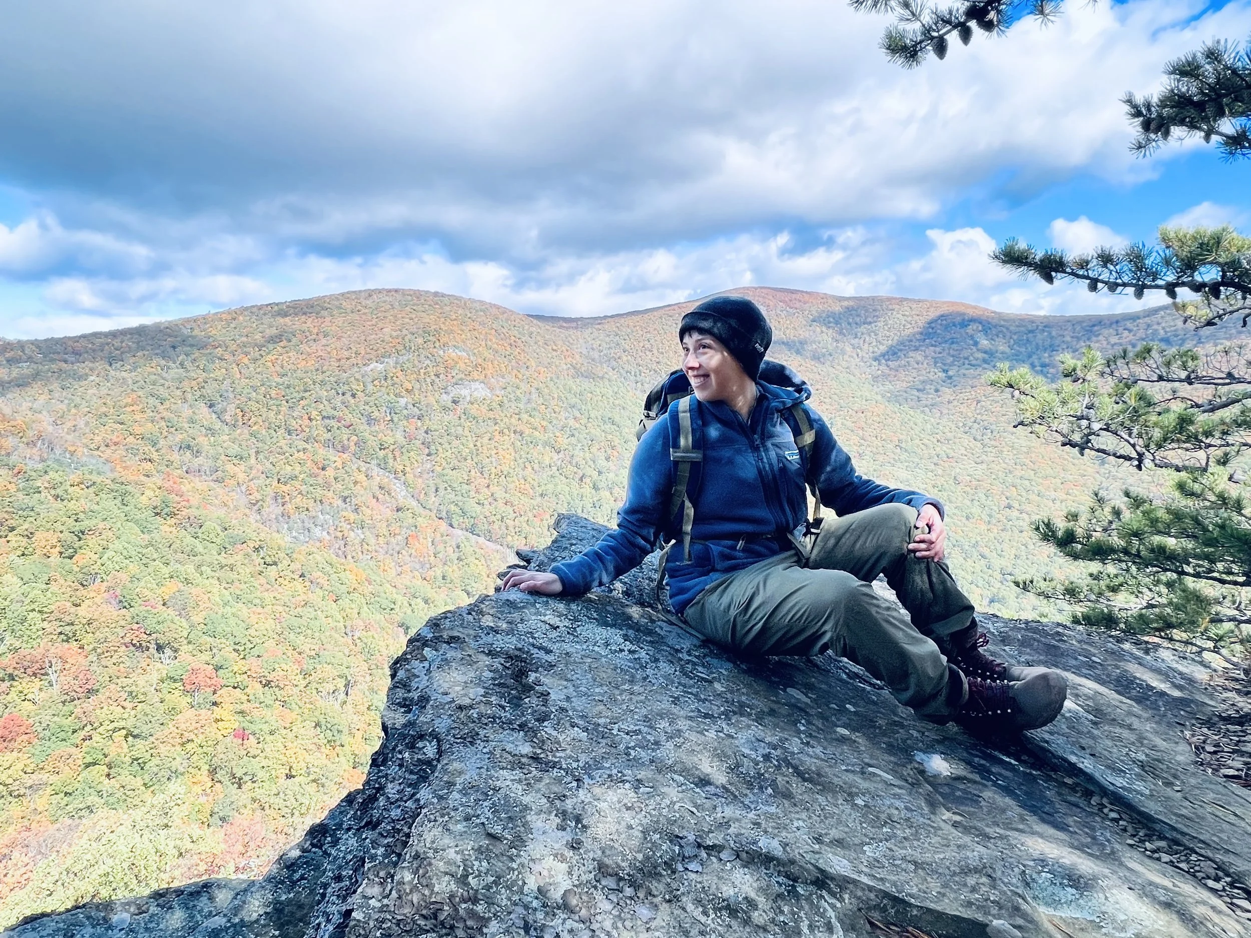 A woman in outdoor hiking gear sitting on a large rock overlooking a colorful fall forested mountain landscape.