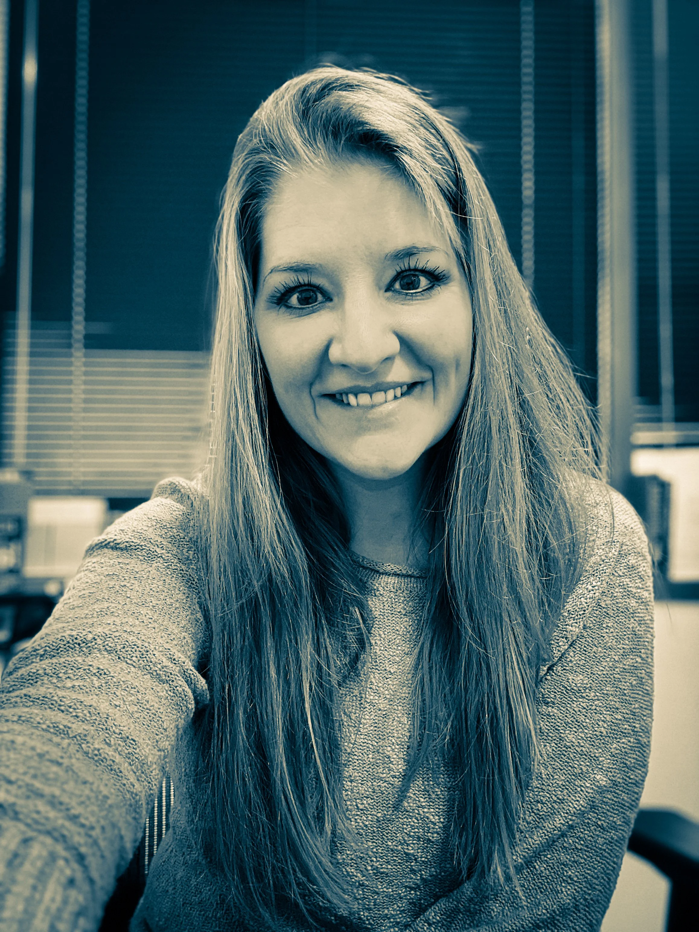 A woman taking a selfie in an office setting with blinds in the background.