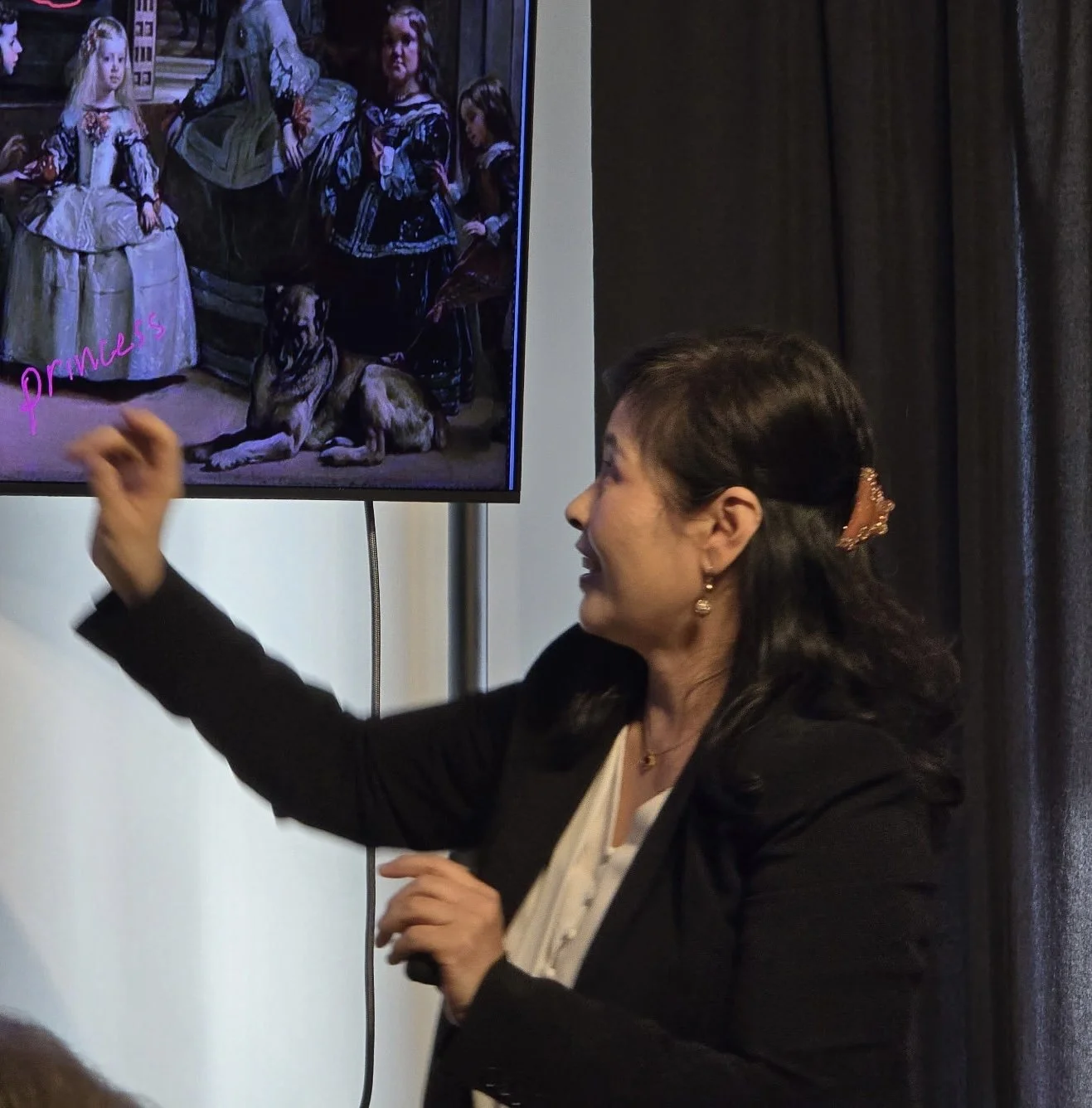 A woman with dark hair and earrings is smiling and gesturing towards a screen displaying artwork of young girls and a dog, with the word 'princess' written on the image.