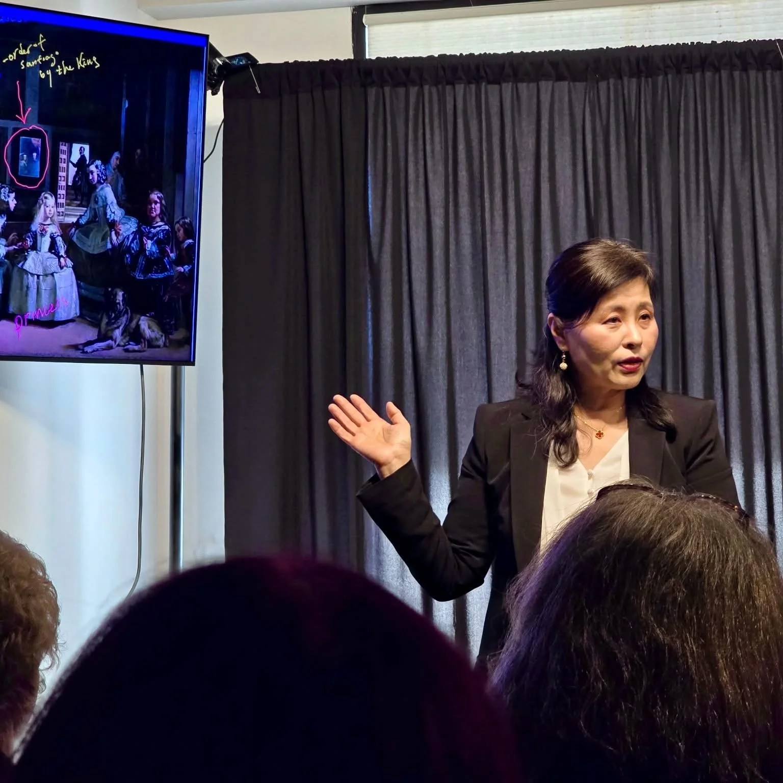 A woman giving a presentation with a black curtain backdrop, pointing towards a screen displaying an image of a theatrical scene with children in costumes, annotated with handwritten notes.