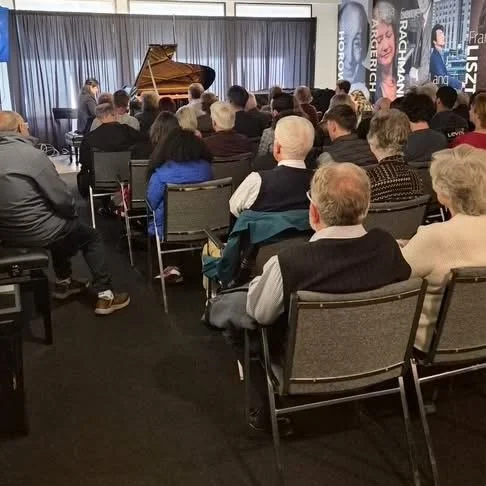 Audience seated in chairs watching a live piano performance on a stage with a grand piano, with banners on the side promoting different shows.