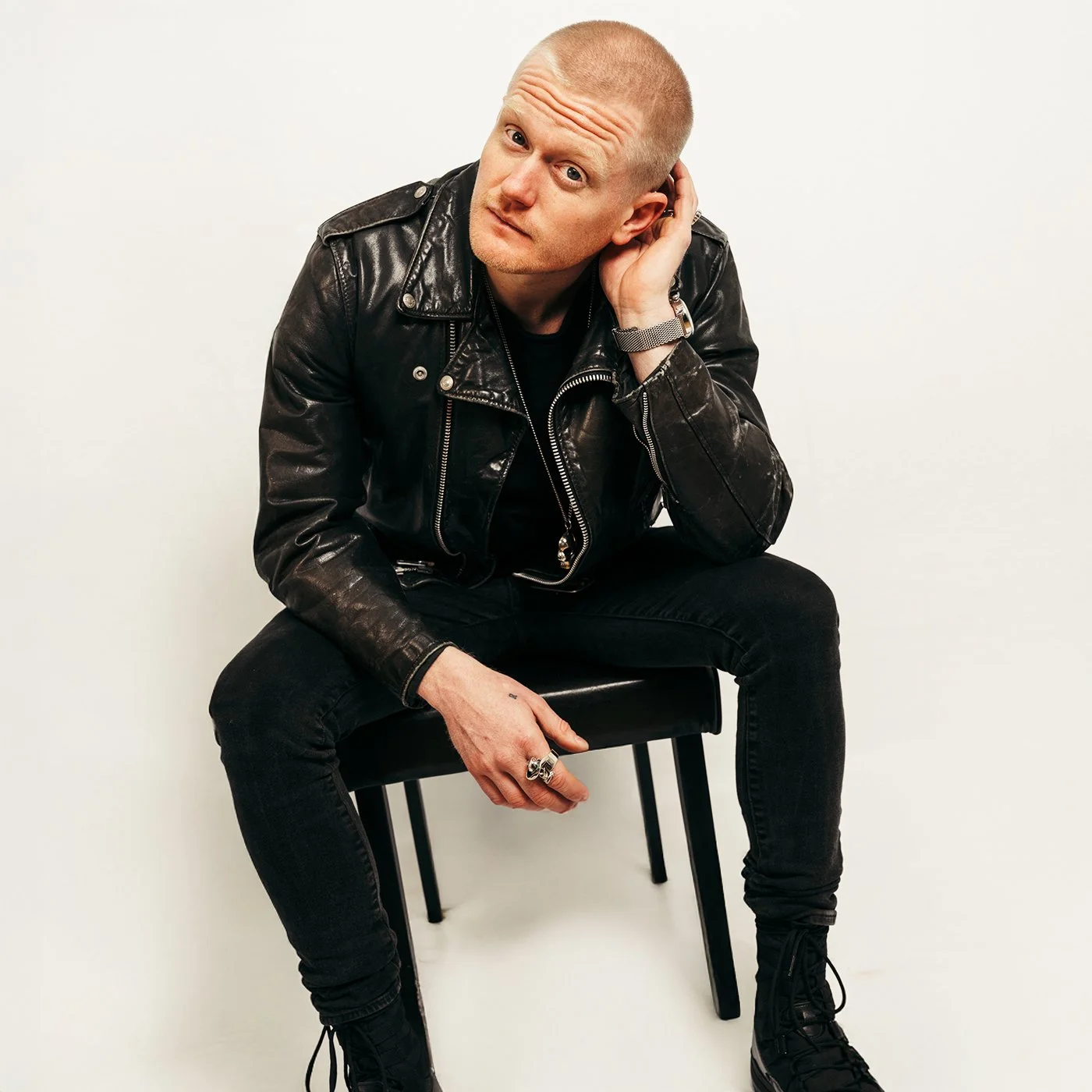 A man with a shaved head wearing a black leather jacket, black pants, and black boots is sitting on a black chair against a plain white background. He is leaning forward with one hand on his knee and the other touching his ear, looking at the camera with a curious expression.