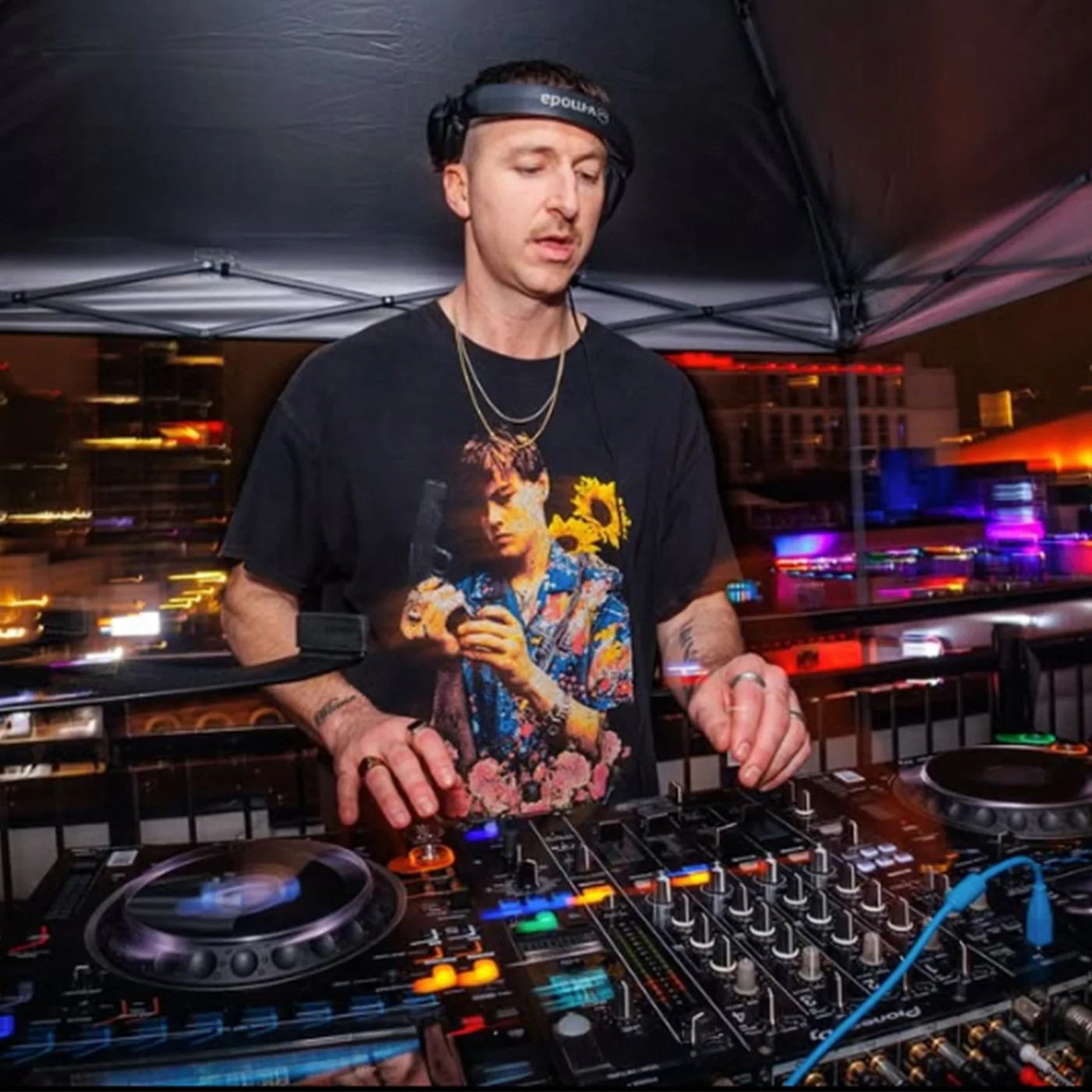 A DJ wearing headphones and a graphic t-shirt with a woman holding a gun is performing at a nightclub with colorful city lights in the background.