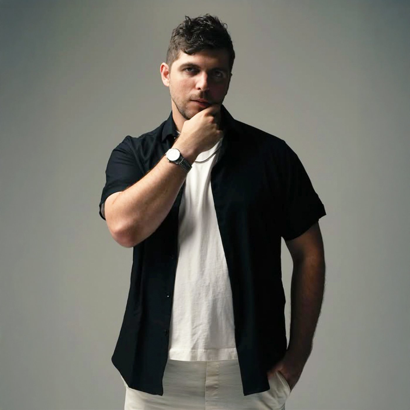 Young man in black shirt and white pants, posing with hand on chin, gray background.