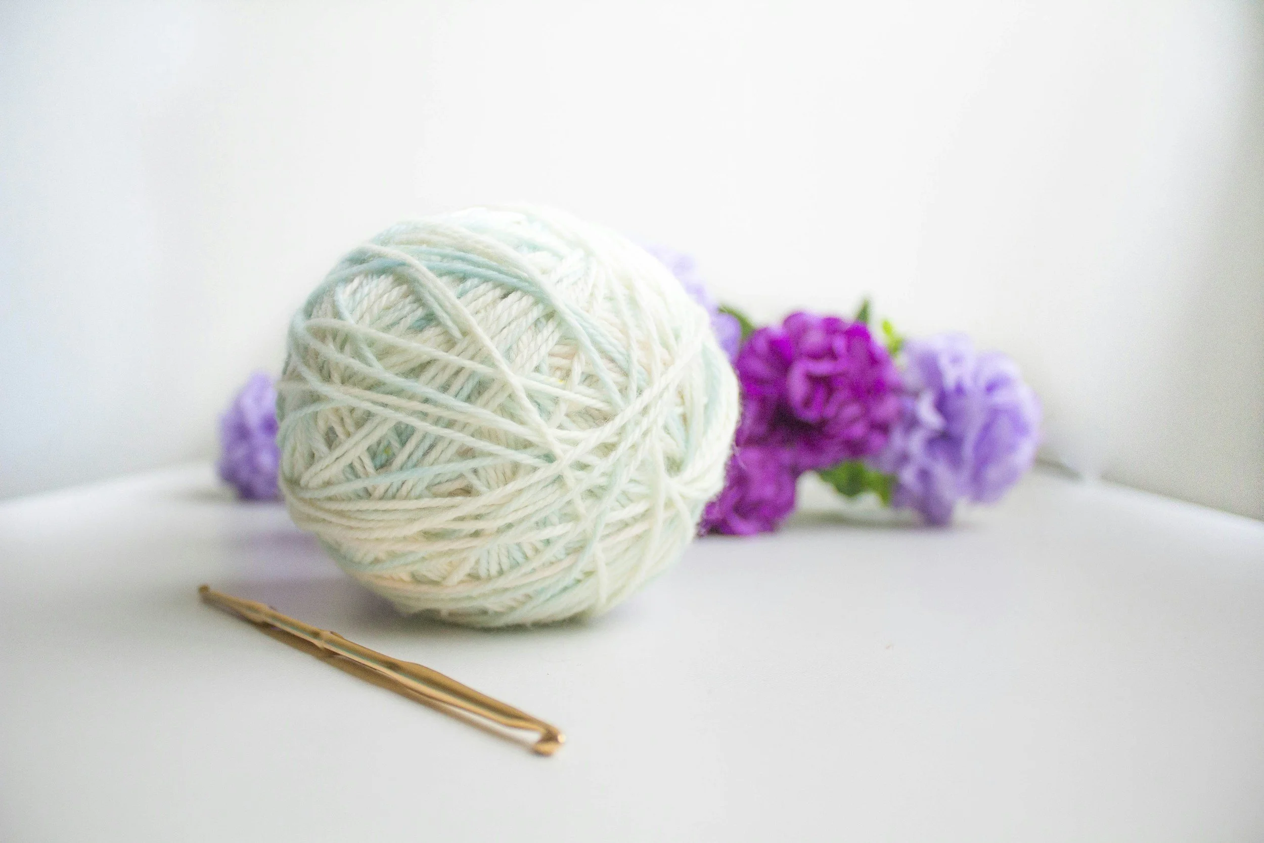 A ball of white yarn, a gold hairpin, and purple lilac flowers on a white surface.