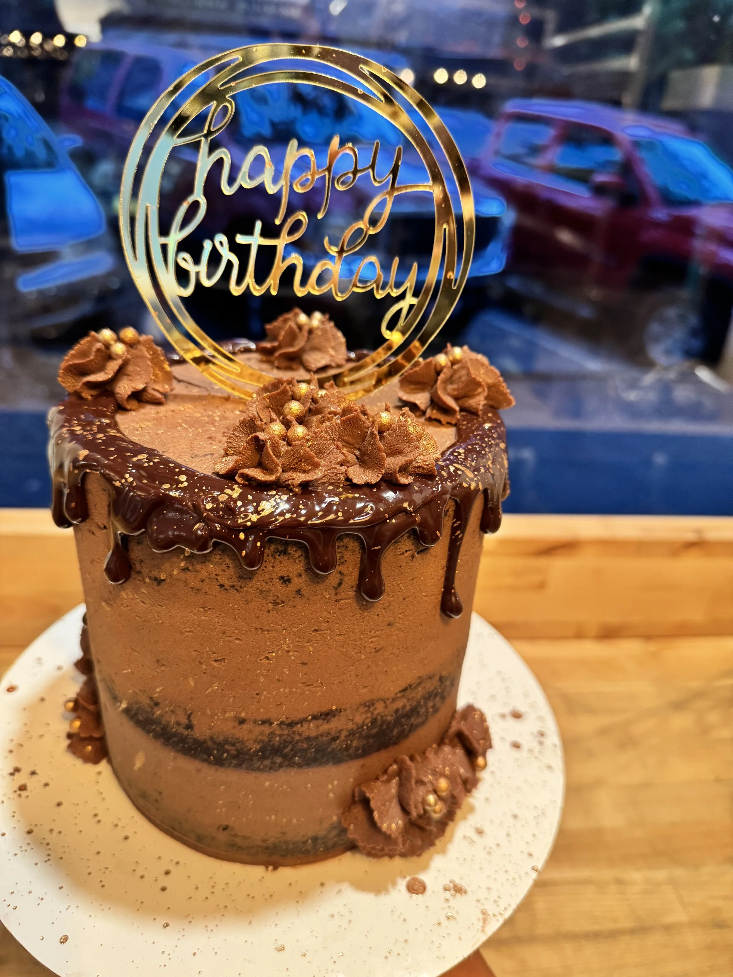 A chocolate birthday cake decorated with chocolate frosting, chocolate drizzle, and chocolate flower decorations, topped with a gold 'happy birthday' sign, on a white cake board with a wooden surface underneath.