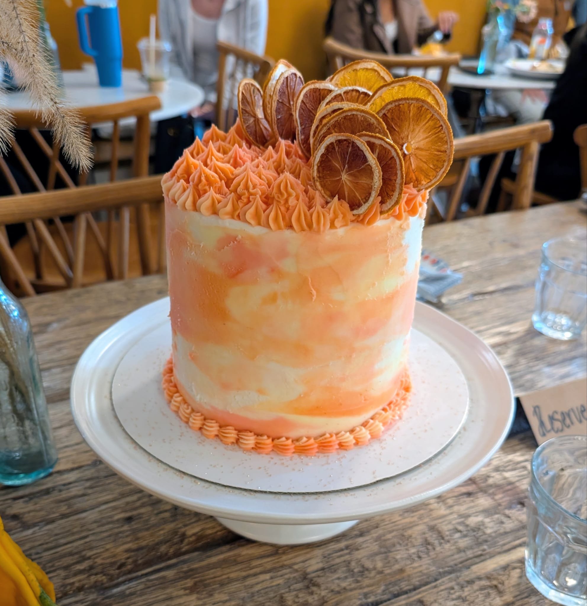 A tall, marble-effect cake with orange and white hues decorated with piped orange frosting and dried orange slices on top, placed on a white cake stand.