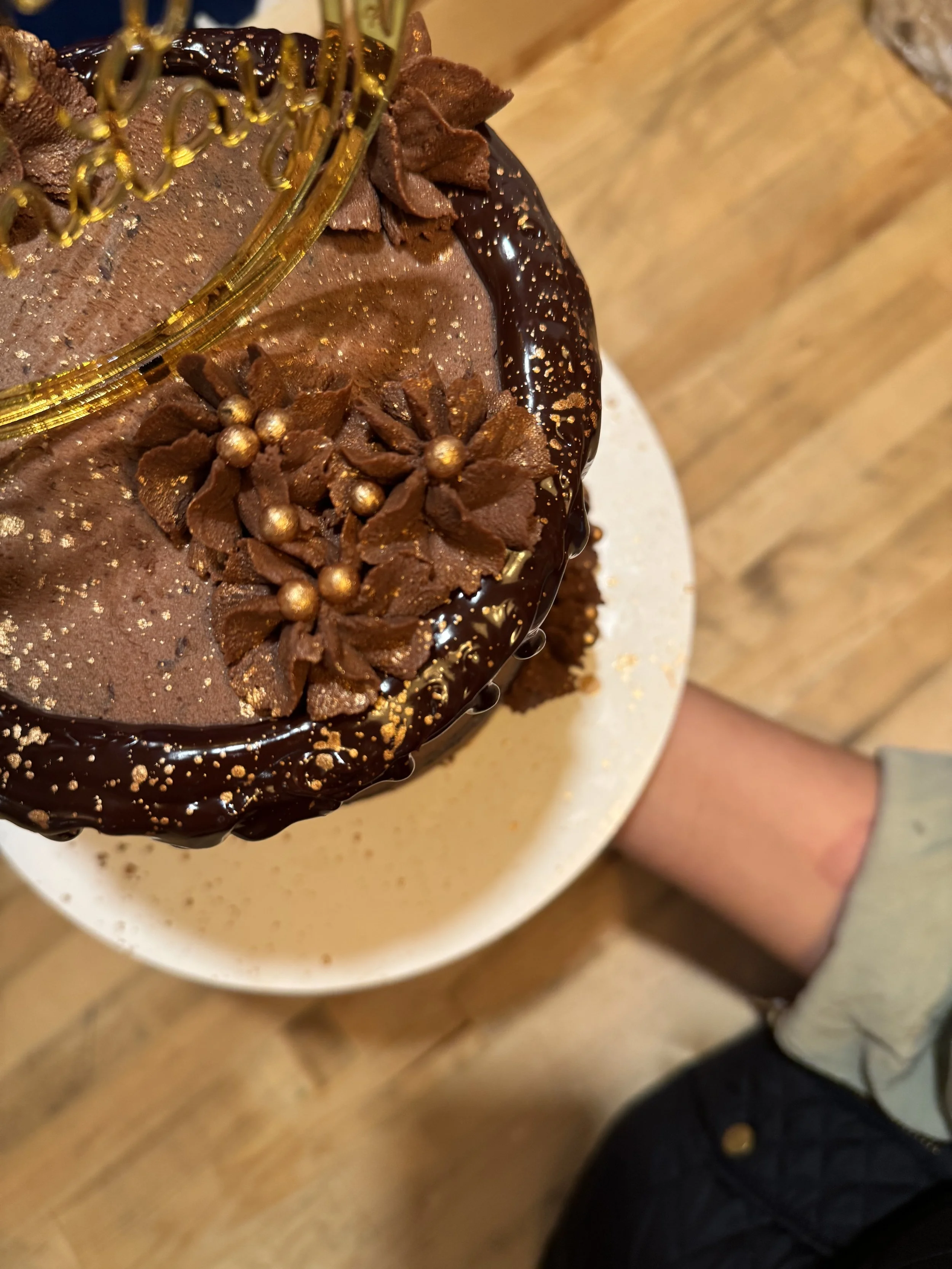 Close-up of a chocolate cake decorated with chocolate flowers, gold sprinkles, and a gold wire decorative piece on top, with a person's arm visible holding the cake.