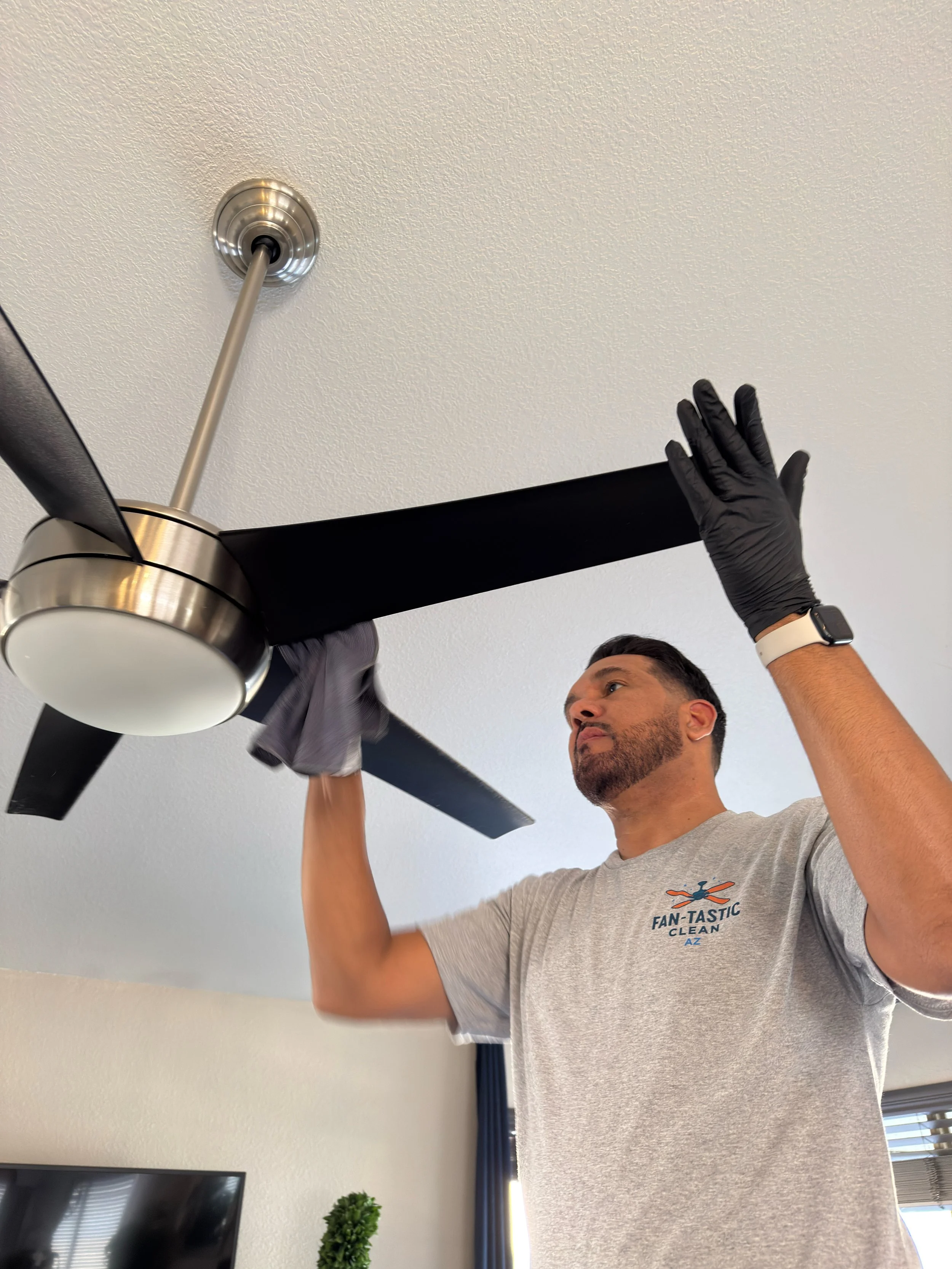 Man cleaning ceiling fan with cloth while wearing black gloves, gray t-shirt, and smartwatch.