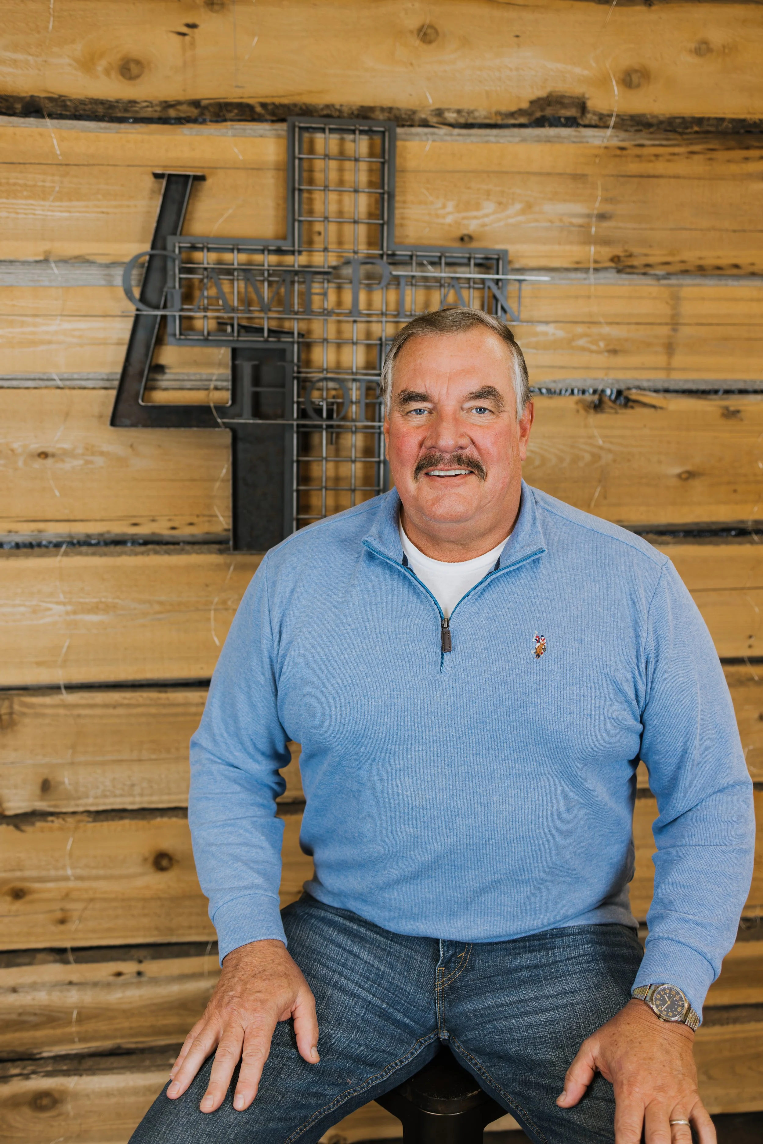 A man with a mustache and short blond hair wearing a dark gray long-sleeve shirt with the words 'Game Plan for Hope' printed on it, standing in an industrial or warehouse setting.