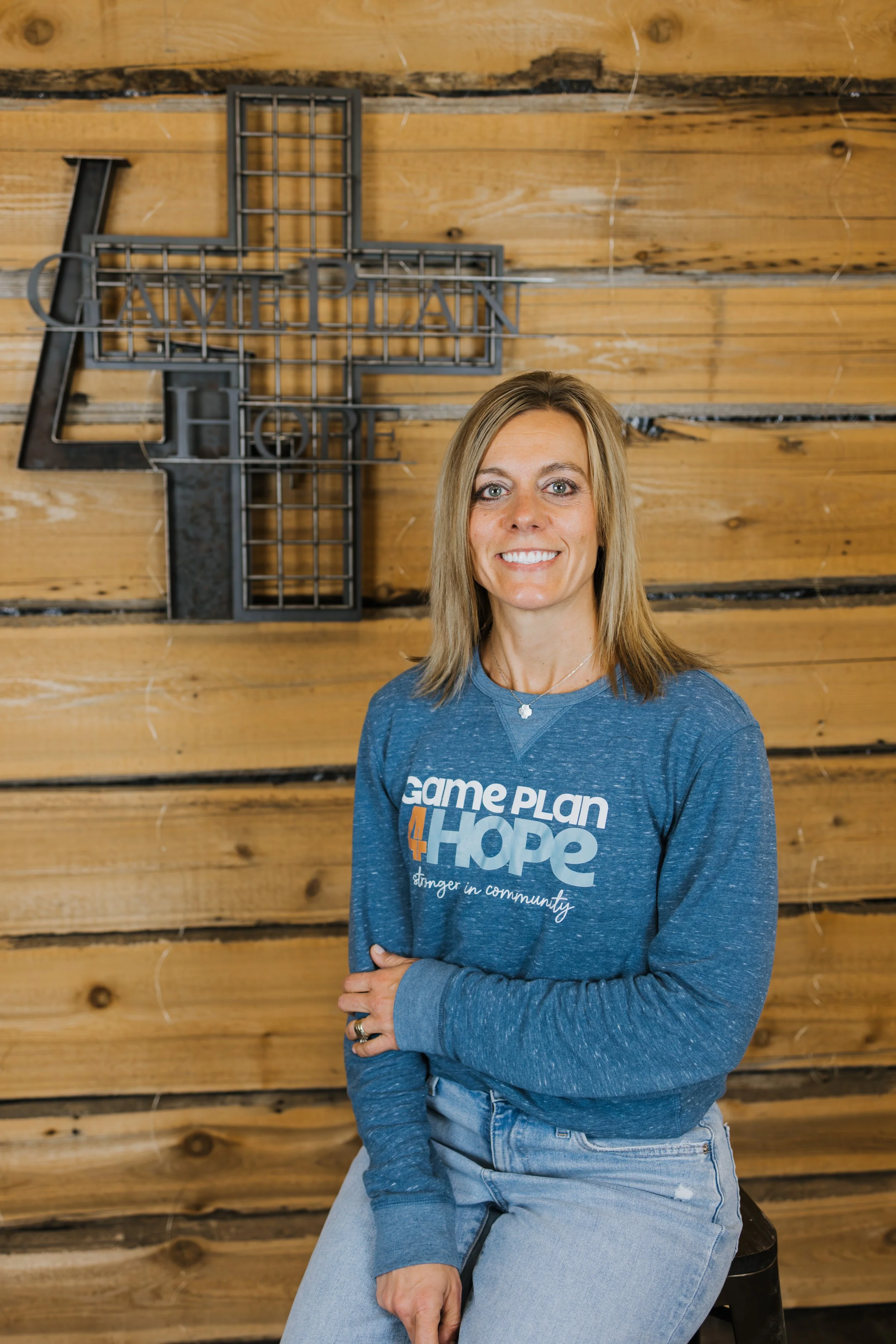 A woman with blonde hair wearing a denim jacket and a white t-shirt, smiling, standing in an indoor workshop or studio with shelves and equipment in the background.