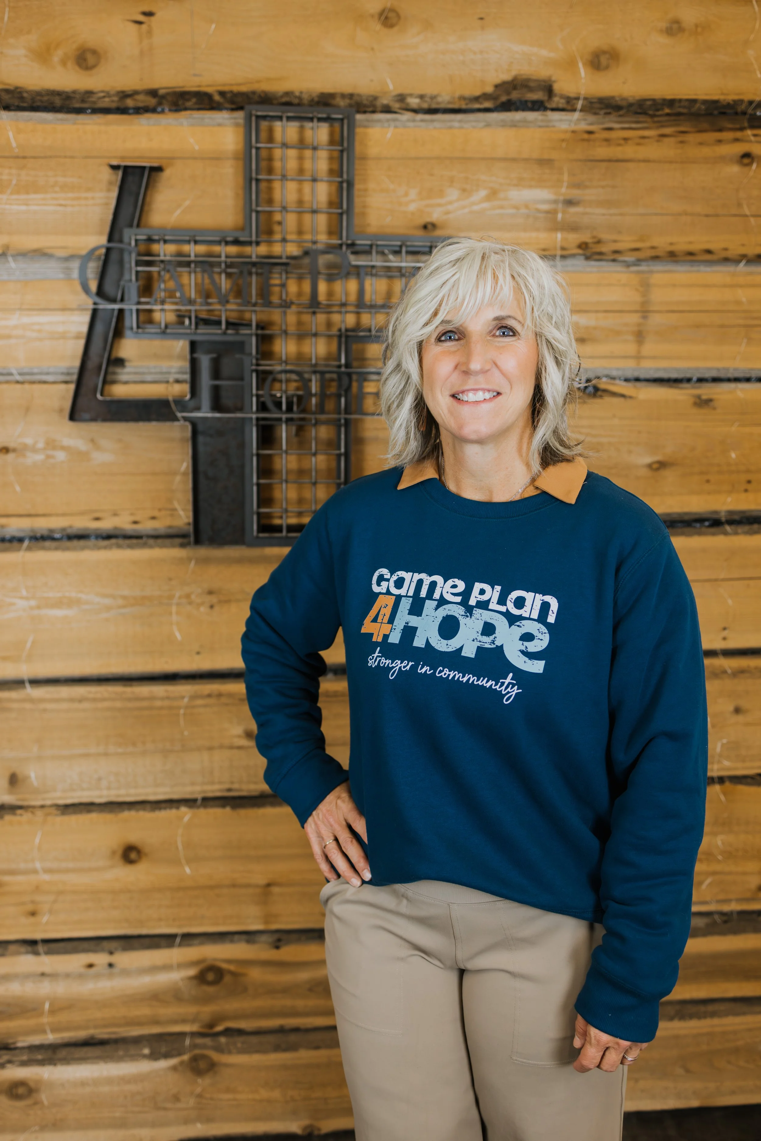 A smiling woman with short gray hair, wearing a black t-shirt with white text, layered with a black and beige cardigan, and silver jewelry, standing in an indoor setting with shelves and equipment in the background.
