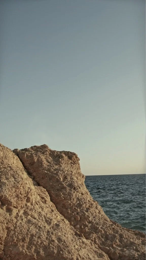 Large beige rocks near the ocean with a clear sky in the background.