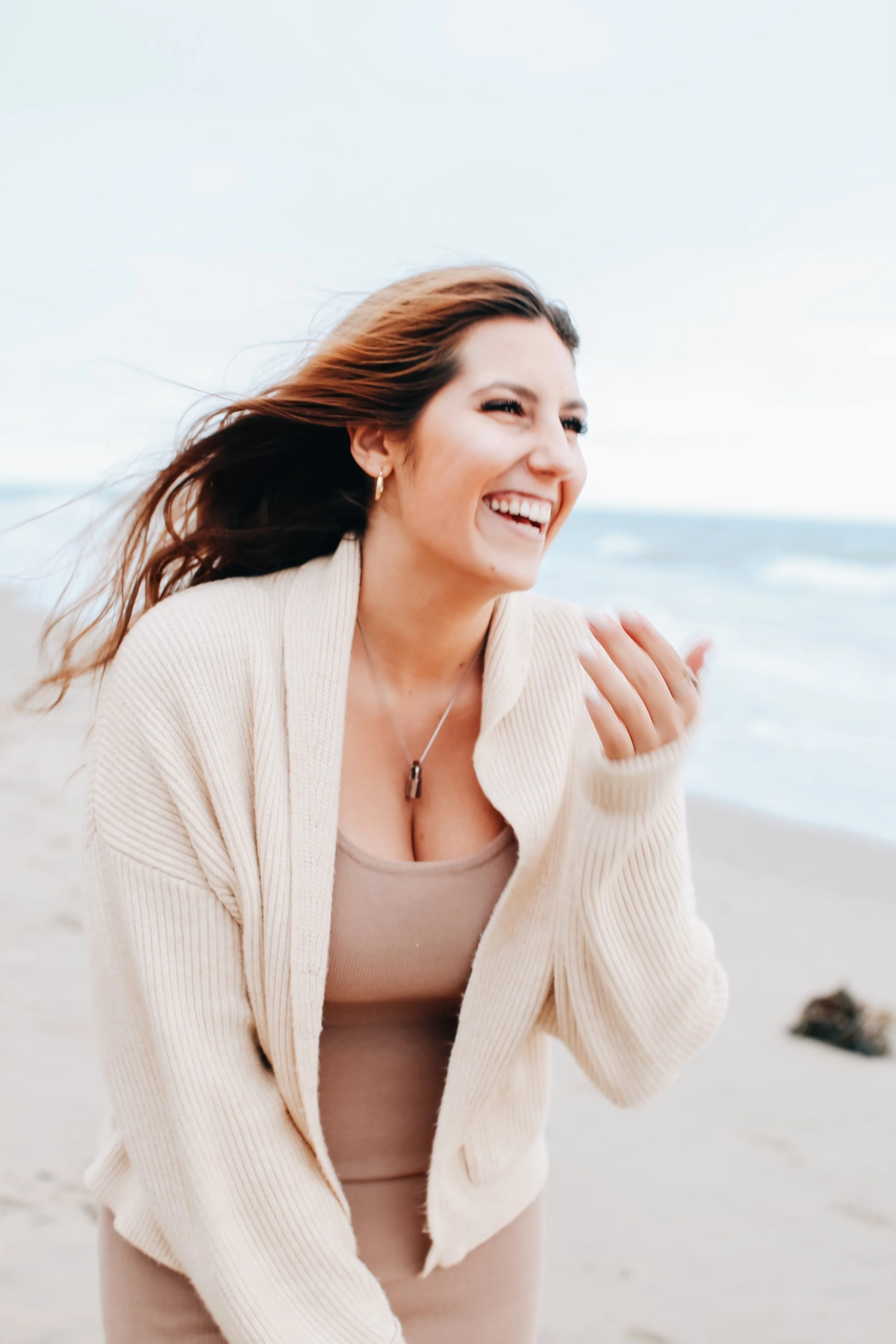 A woman with brown hair smiling at the beach, wearing a beige cardigan and tank top, with a blurred ocean background.