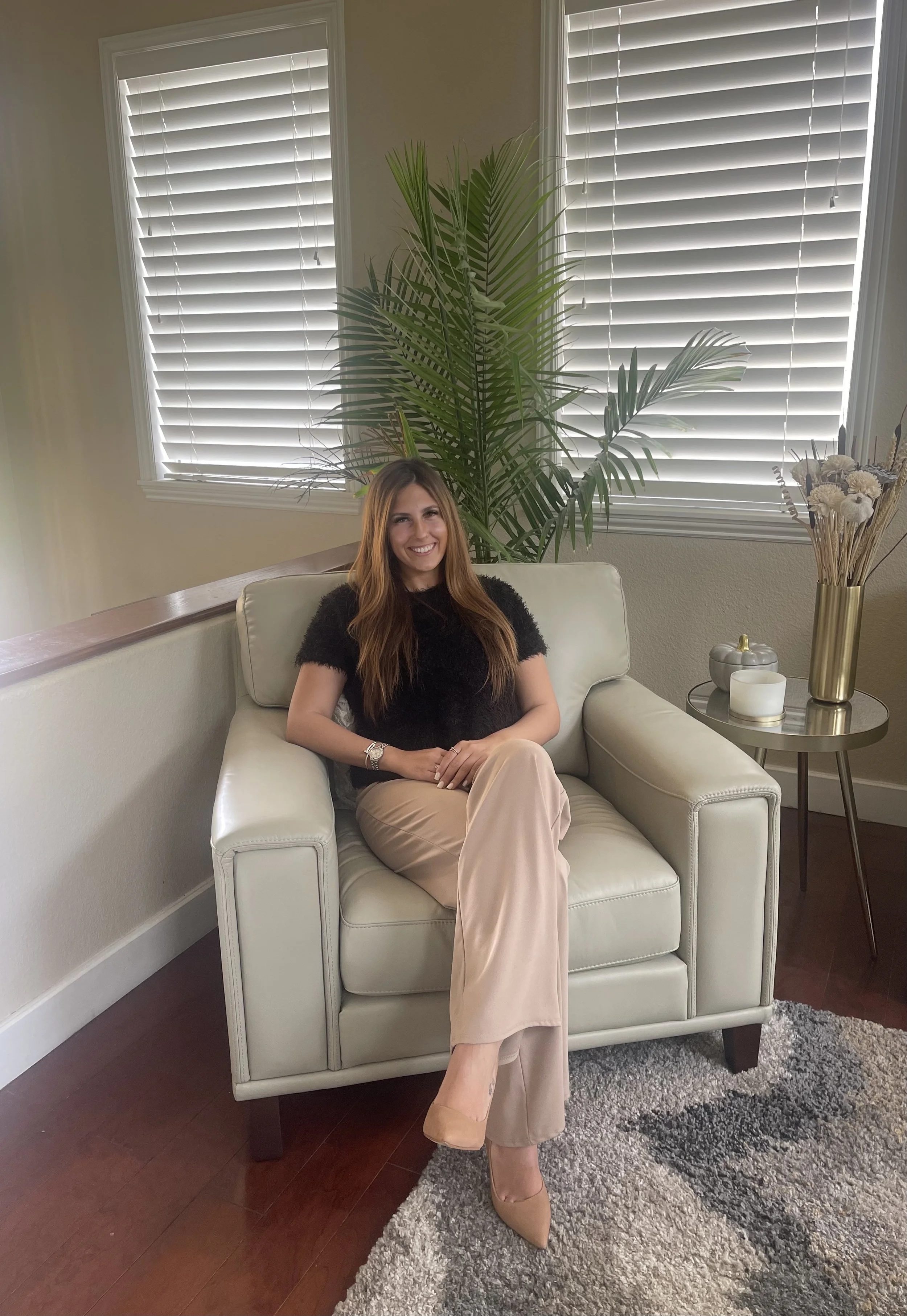 Woman with long brown hair sitting on a white sofa in a living room, smiling, wearing a black top and beige pants, with large green houseplant and window with white blinds behind her, side table with decorative items to her right.
