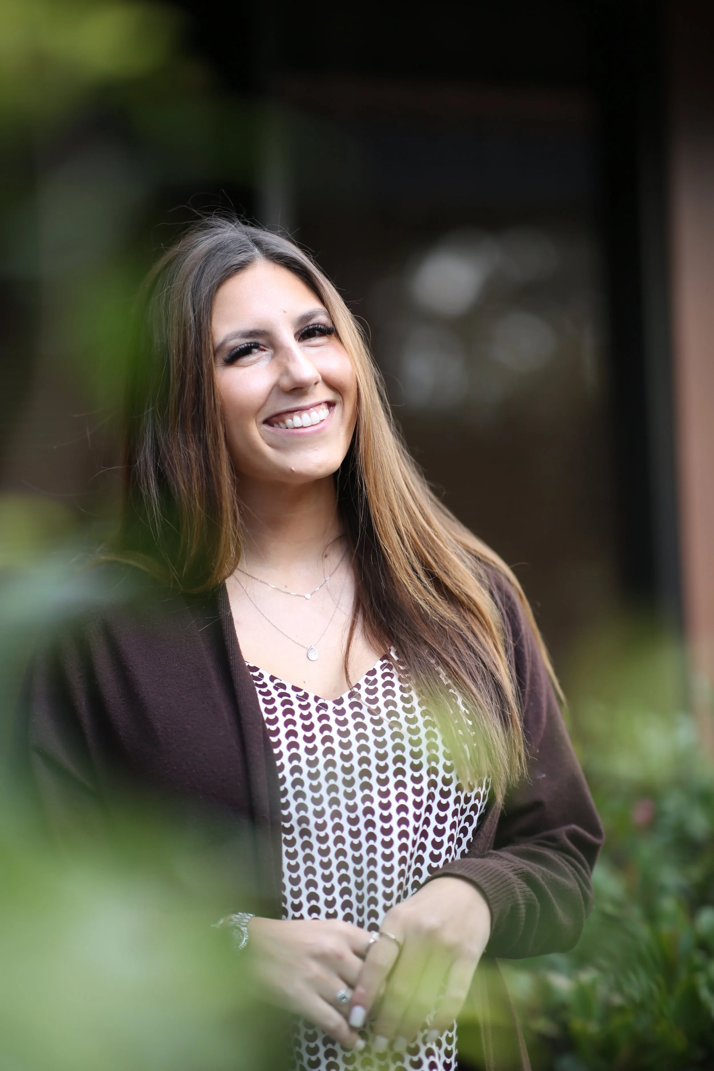 A young woman with long, light brown hair smiling outdoors, wearing a patterned dress, a brown cardigan, and jewelry, with greenery blurred in the foreground.