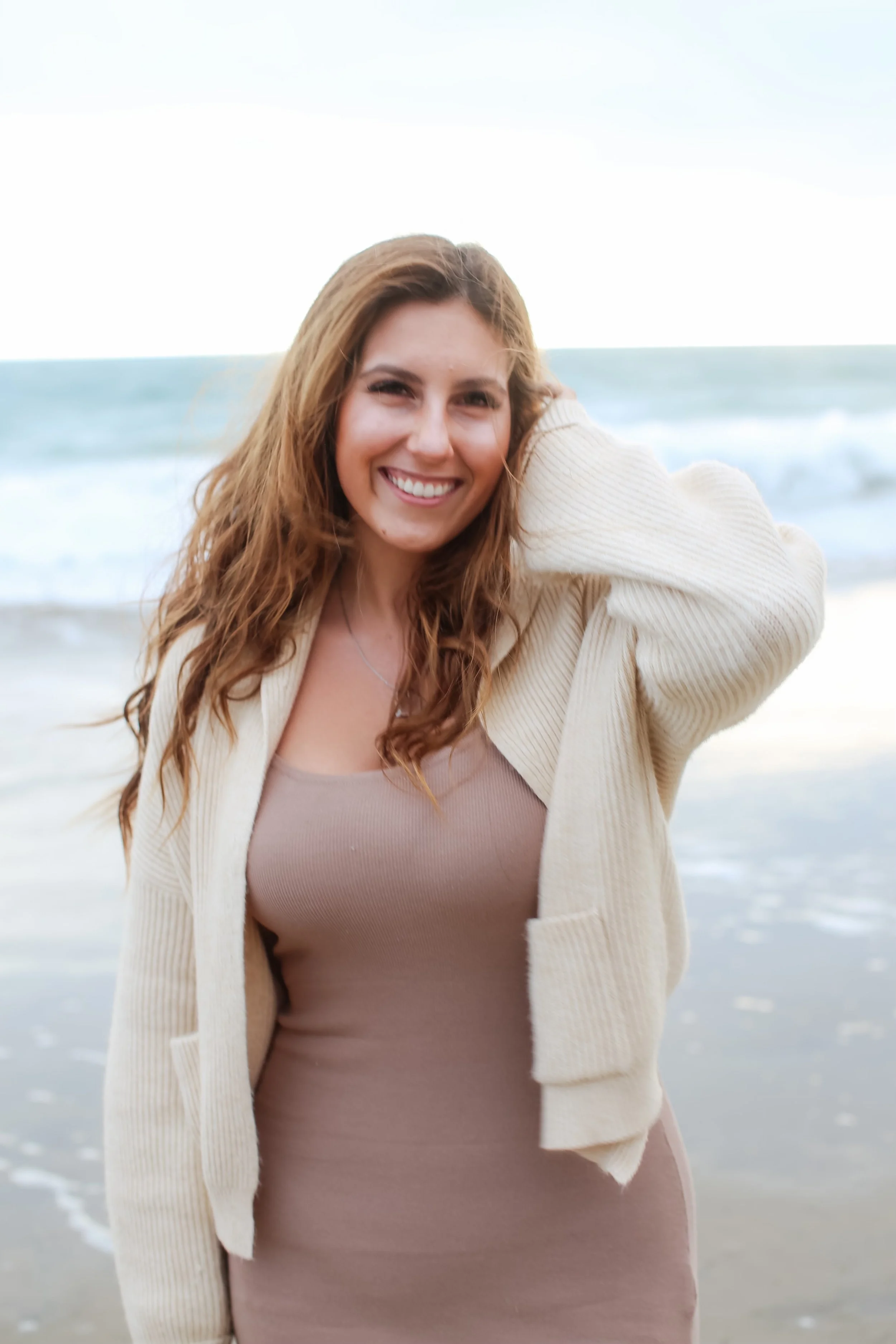 A young woman with long, wavy brown hair smiling while standing on a beach with ocean waves in the background. She is wearing a beige cardigan and a mauve dress.
