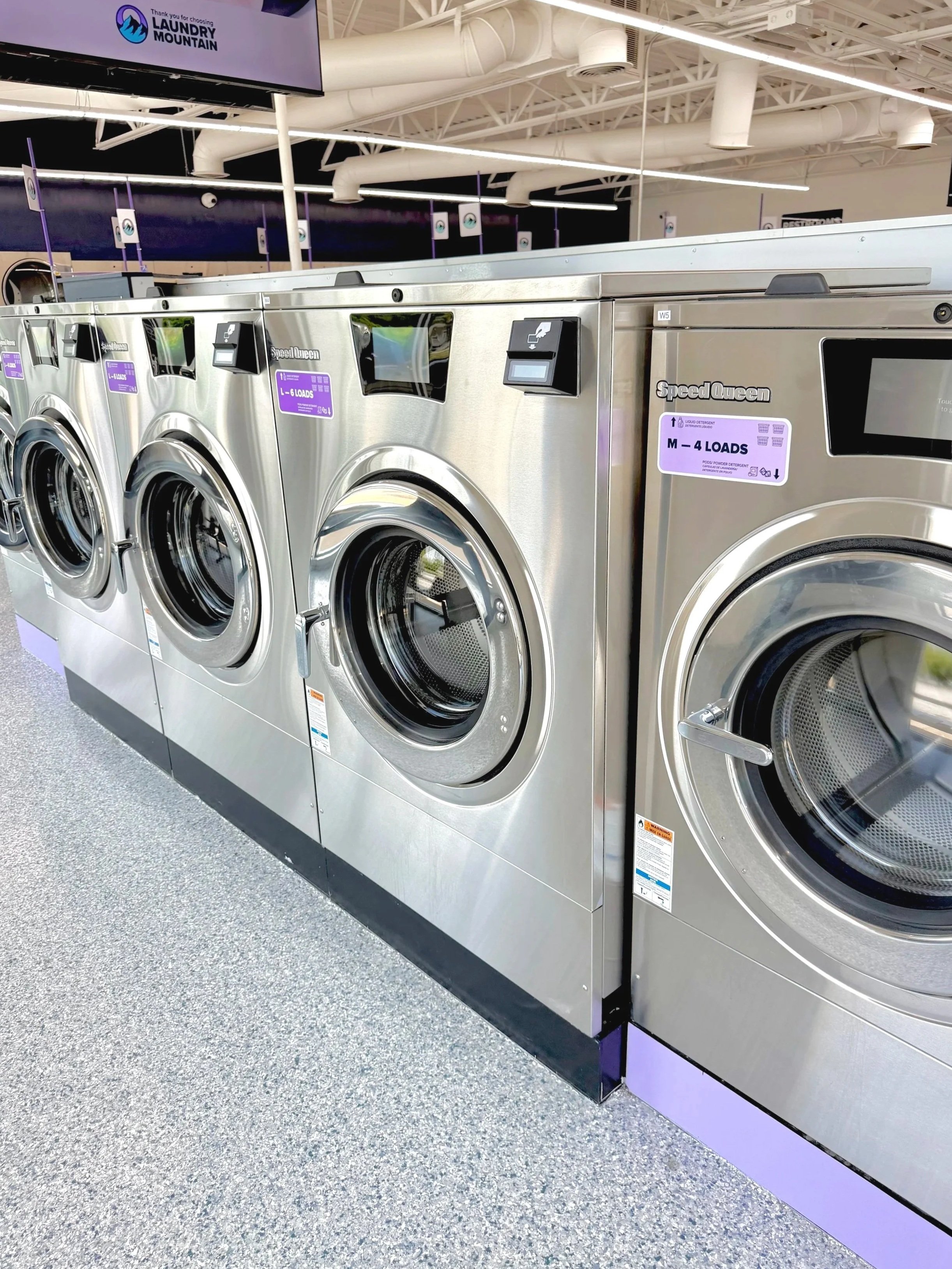 Multiple stainless steel washing machines in a laundromat, with purple labels indicating load capacities, and a overhead screen displaying a laundry service advertisement.