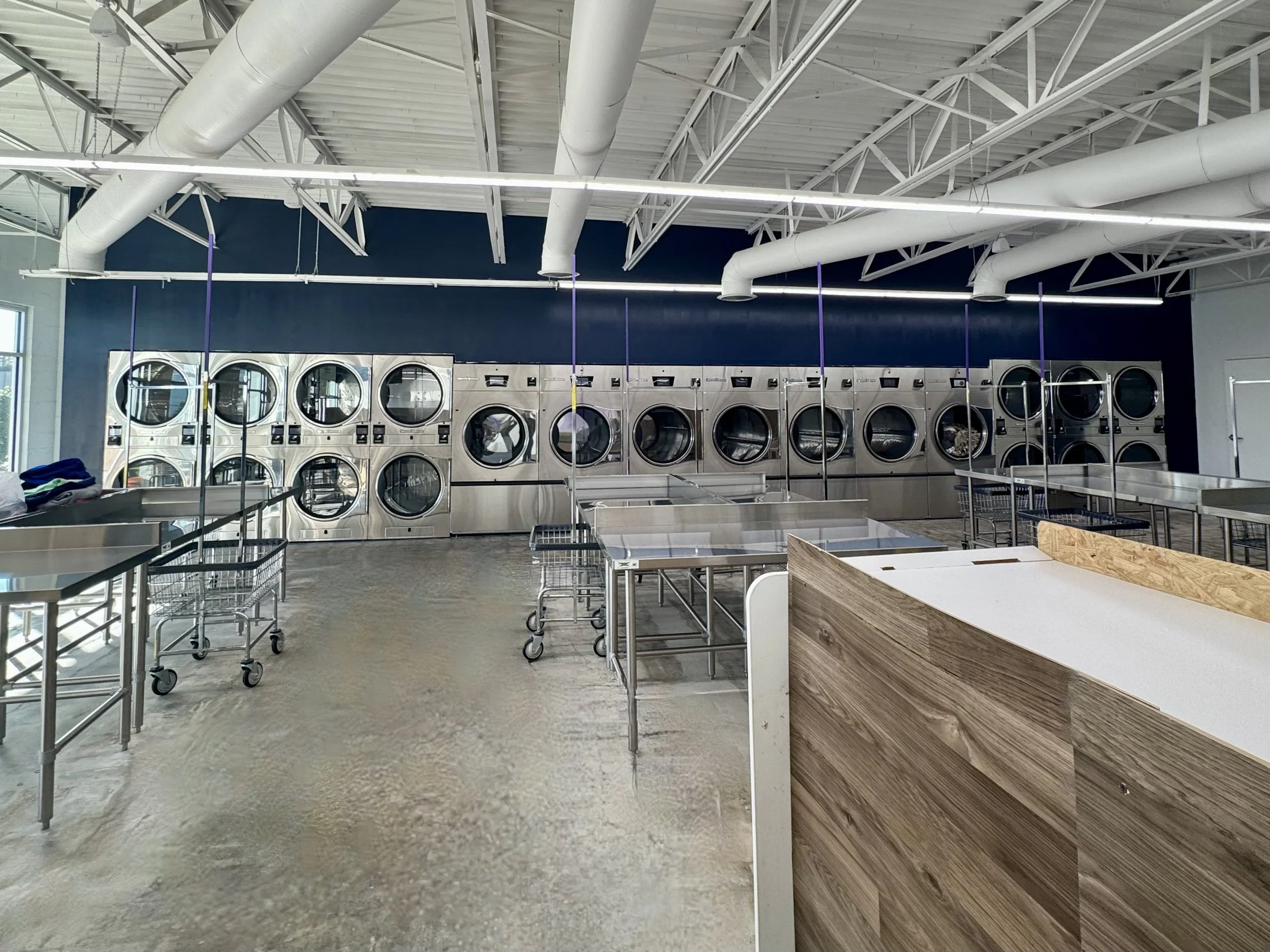 A row of four commercial laundry washing machines in a laundromat with windows in the background.