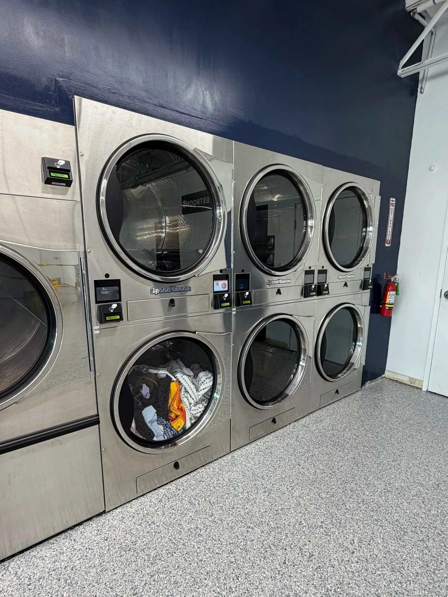 Laundry room with six front-loading washing machines, one filled with clothes, a fire extinguisher on the wall.