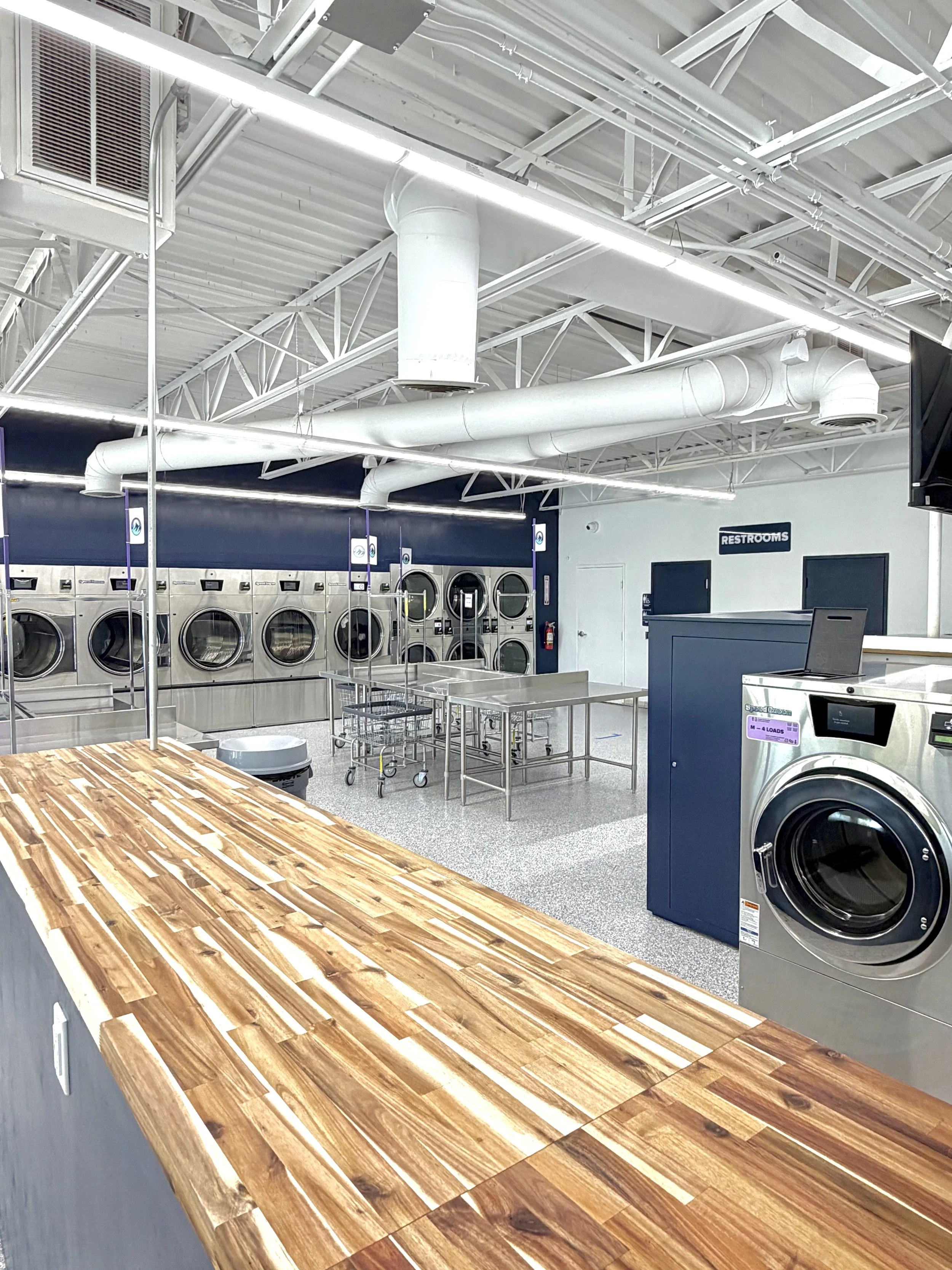 A row of four commercial laundry washing machines in a laundromat with windows in the background.
