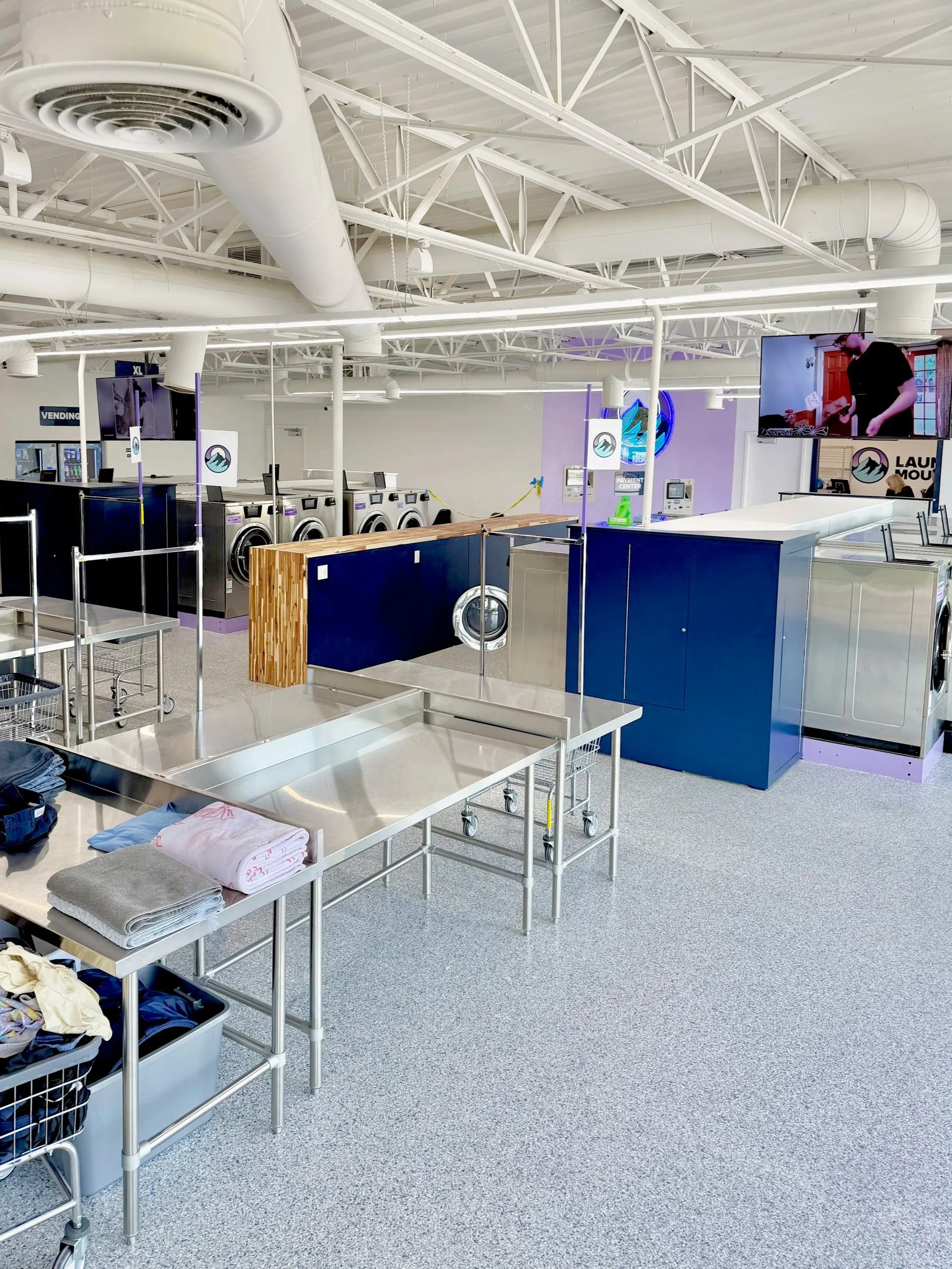 Laundromat with multiple washing machines, laundry tables, and a television screen.