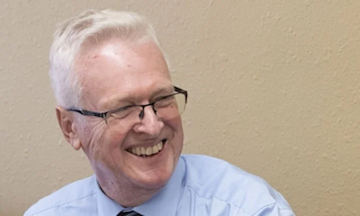 A smiling man with glasses and gray hair wearing a black polo shirt with a logo, sitting on a stone wall outdoors, surrounded by green plants and trees.