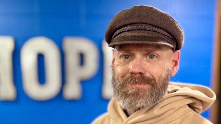 Close-up of a middle-aged man with glasses, blond hair, wearing a gray hoodie and a dark jacket, against a black background.
