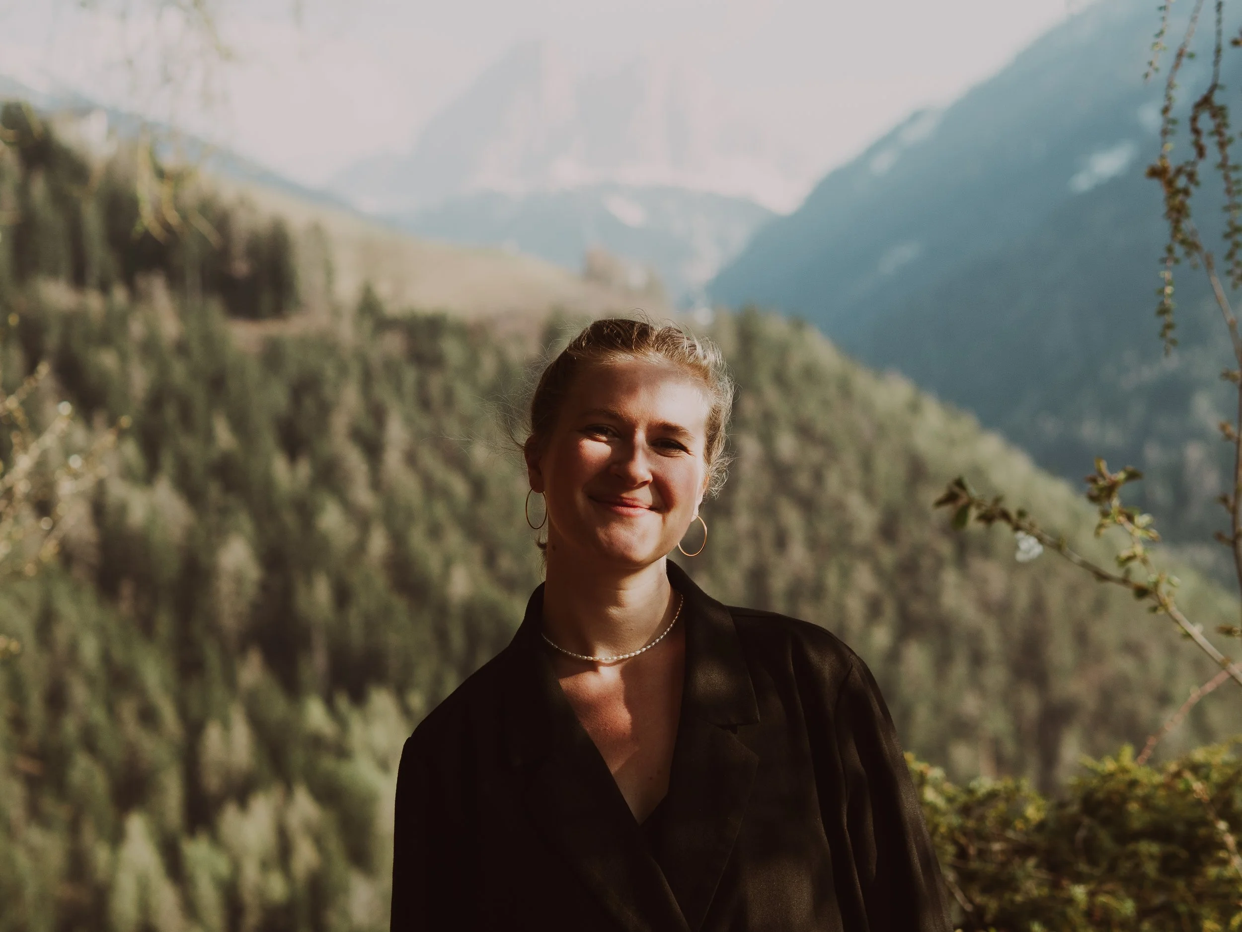 Eine lächelnde Frau mit Ohrringen und Halskette vor einer Berglandschaft mit Wäldern und Bergen im Hintergrund.
