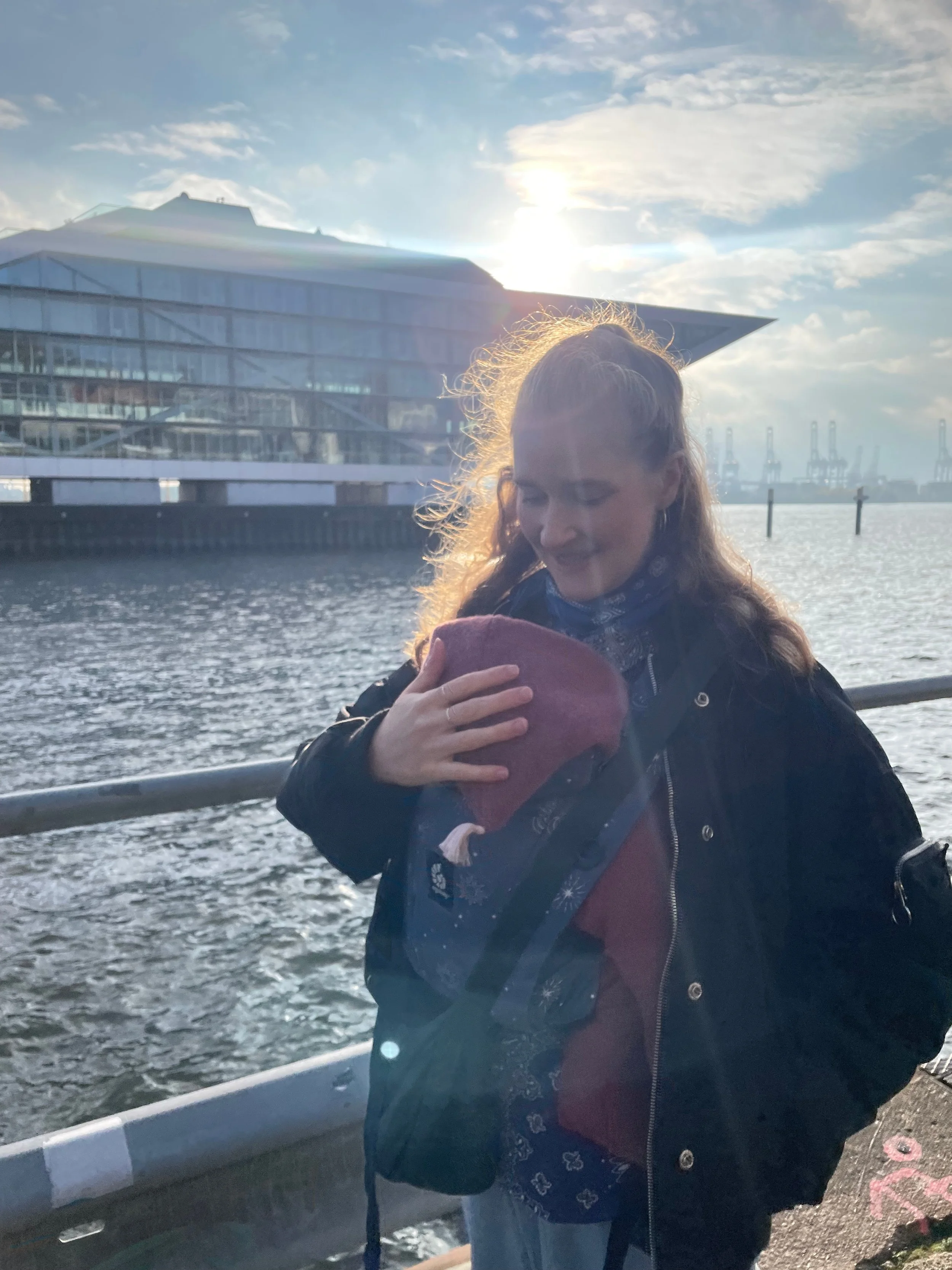 Eine Frau hält ein Baby auf einem Arm, während sie an einer Wasserfront steht, mit moderner Gebäudearchitektur im Hintergrund und der Sonne, die am Himmel untergeht.