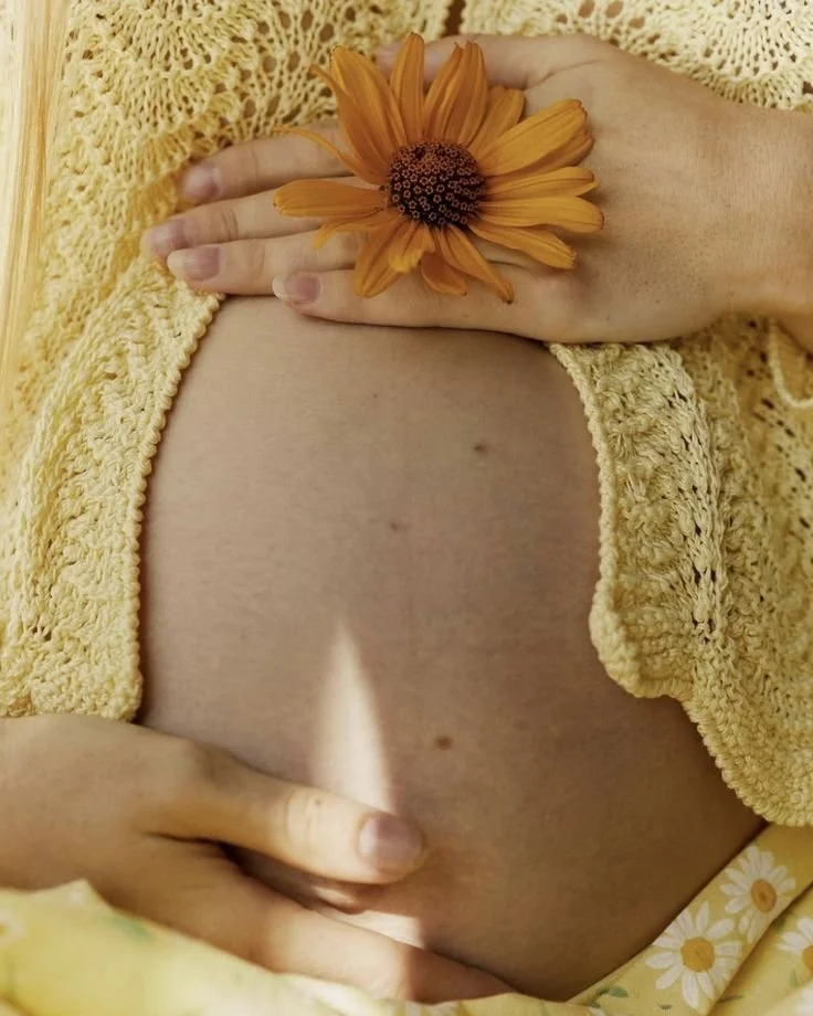 Nahaufnahme eines Bauches mit einer Hand, die eine orange Blume hält, und einer anderen Hand, die den Bauch berührt. Die Person trägt eine gelbe, gestrickte Jacke und sitzt auf einem Stoff mit Blumendesign.