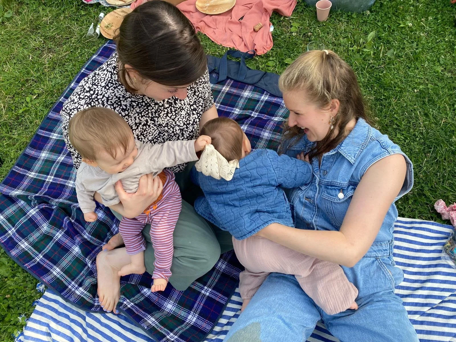 Zwei Frauen spielen mit drei kleinen Kindern auf einer Wiese, sitzen auf Decken, lachen und interagieren miteinander.