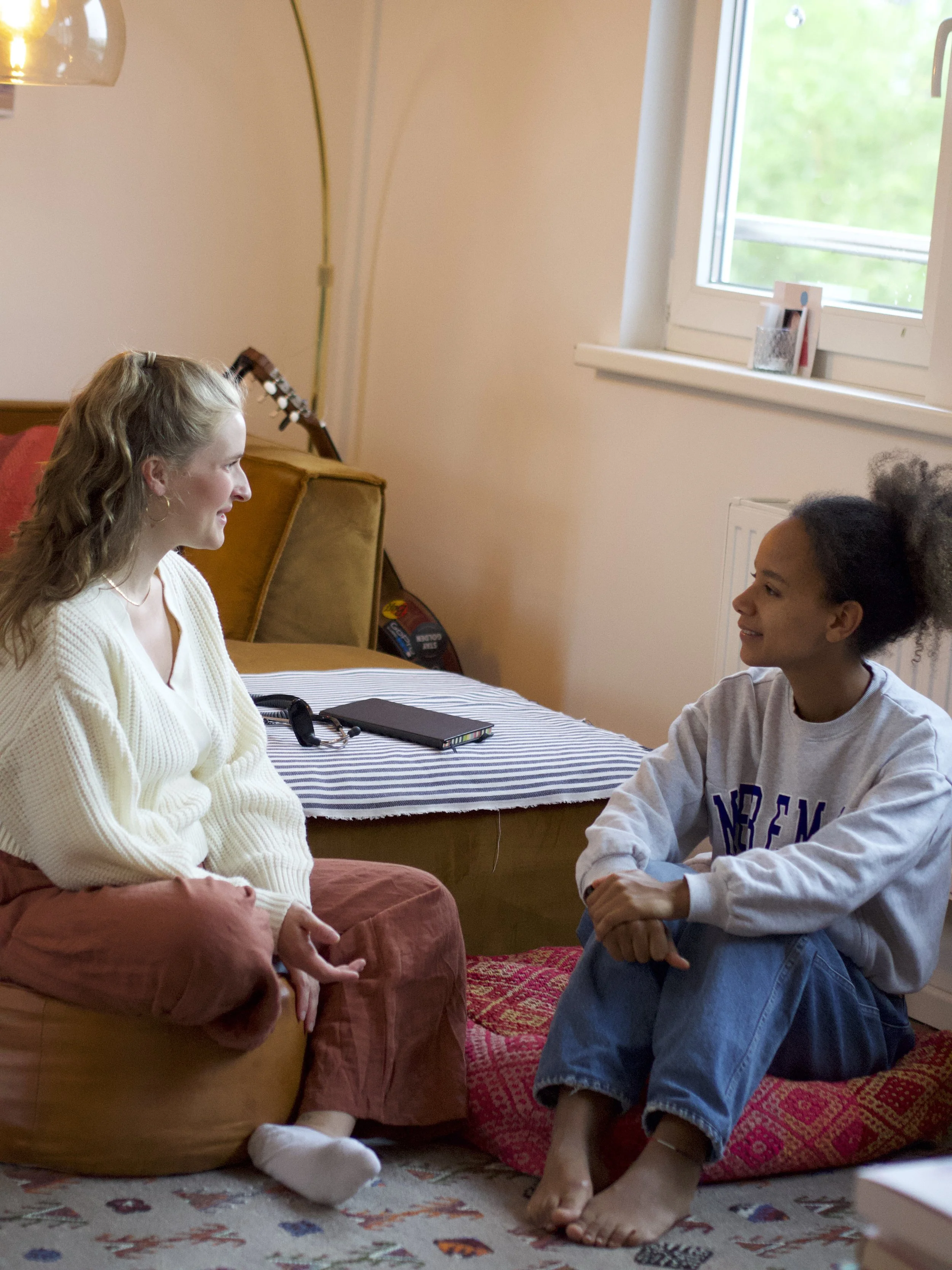 Zwei Frauen sitzen im Wohnzimmer auf Kissen auf dem Boden und unterhalten sich.