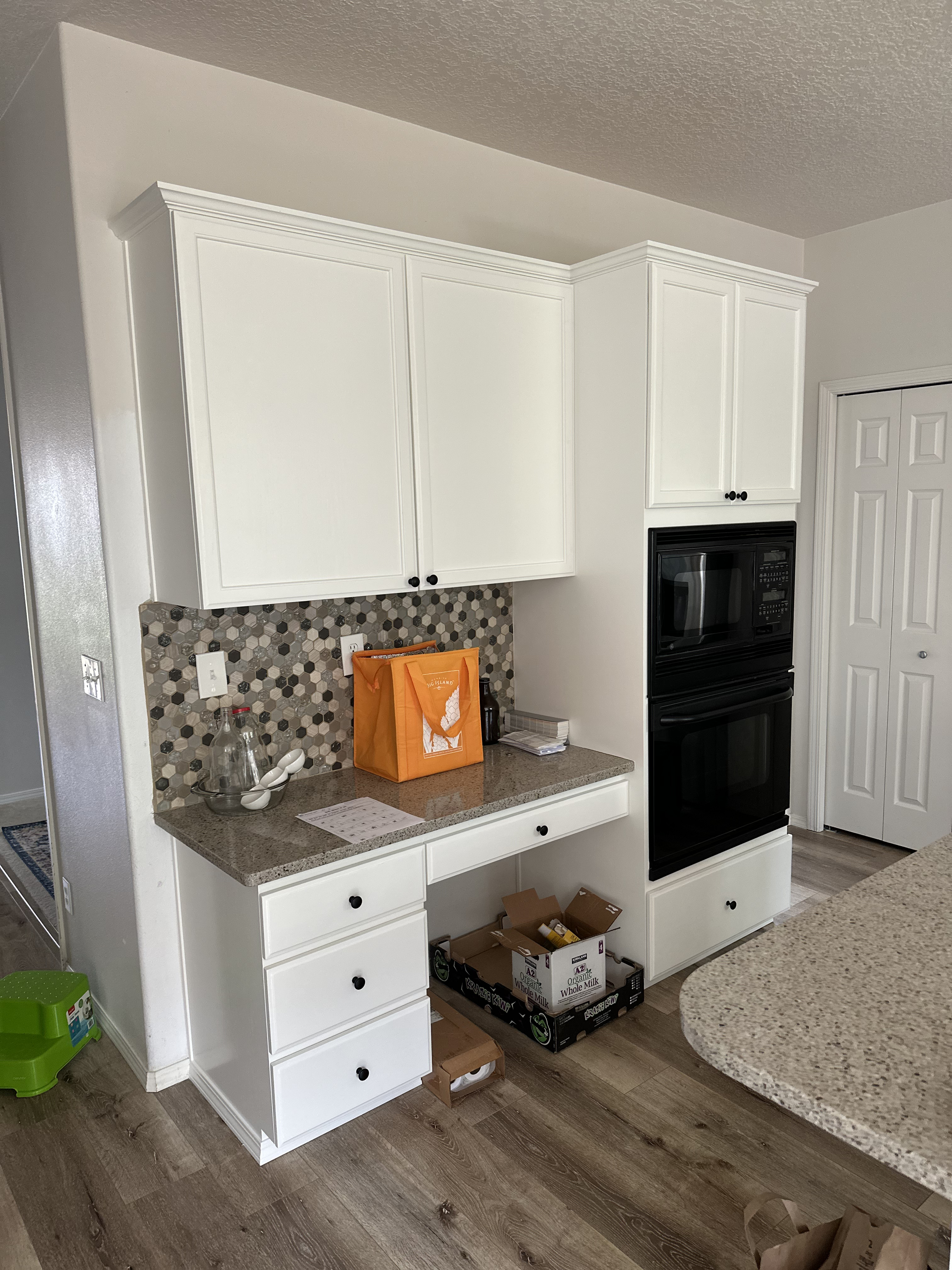 Kitchen corner with white cabinets, a black double oven, a countertop with an orange grocery bag, a glass jar, and some paper; there is a tile backsplash with a honeycomb pattern and a portion of a breakfast bar with granite surface.