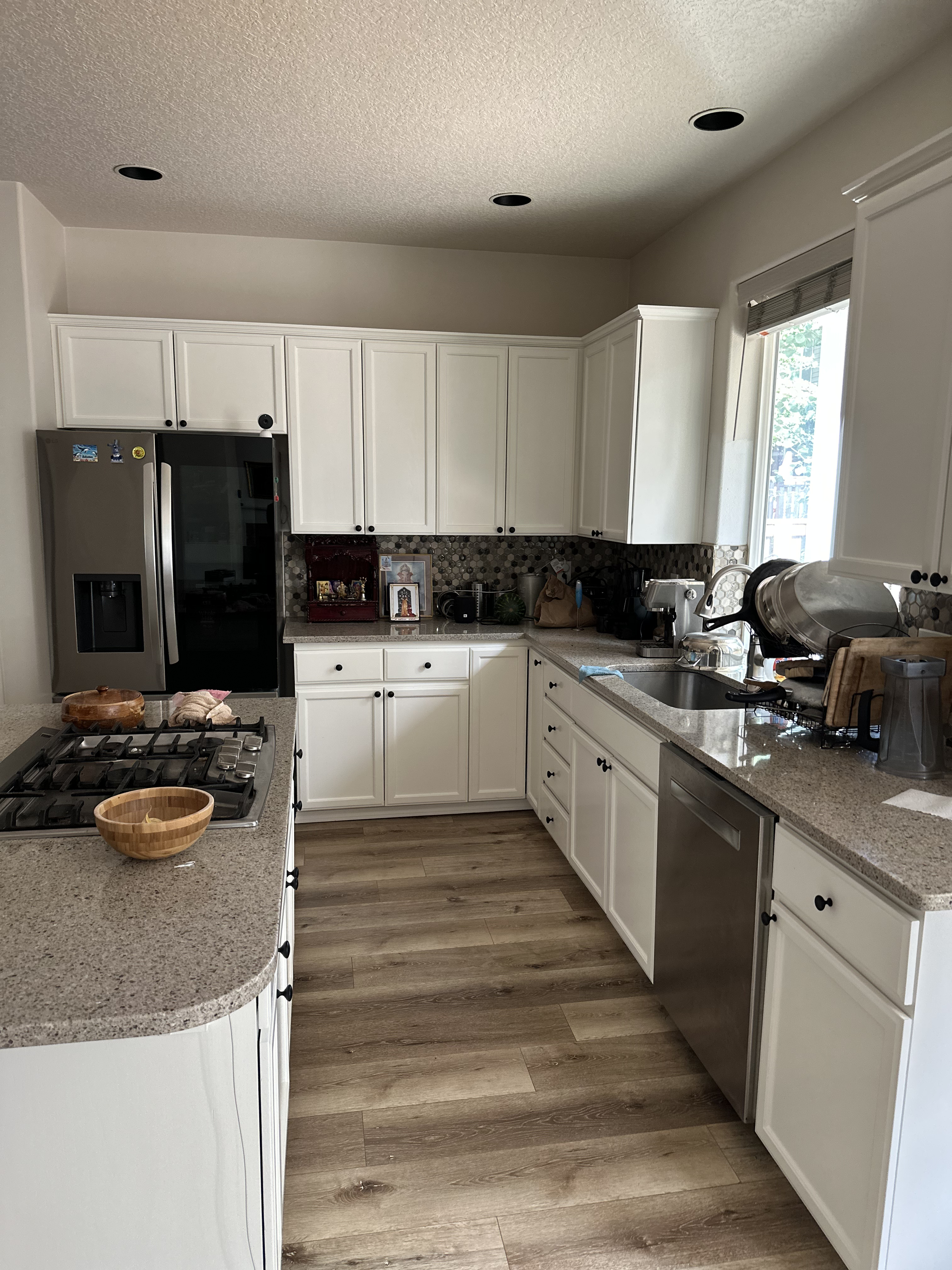 A kitchen with white cabinets, a granite countertop, a stainless steel refrigerator, and various kitchen appliances. A window lets in natural light.