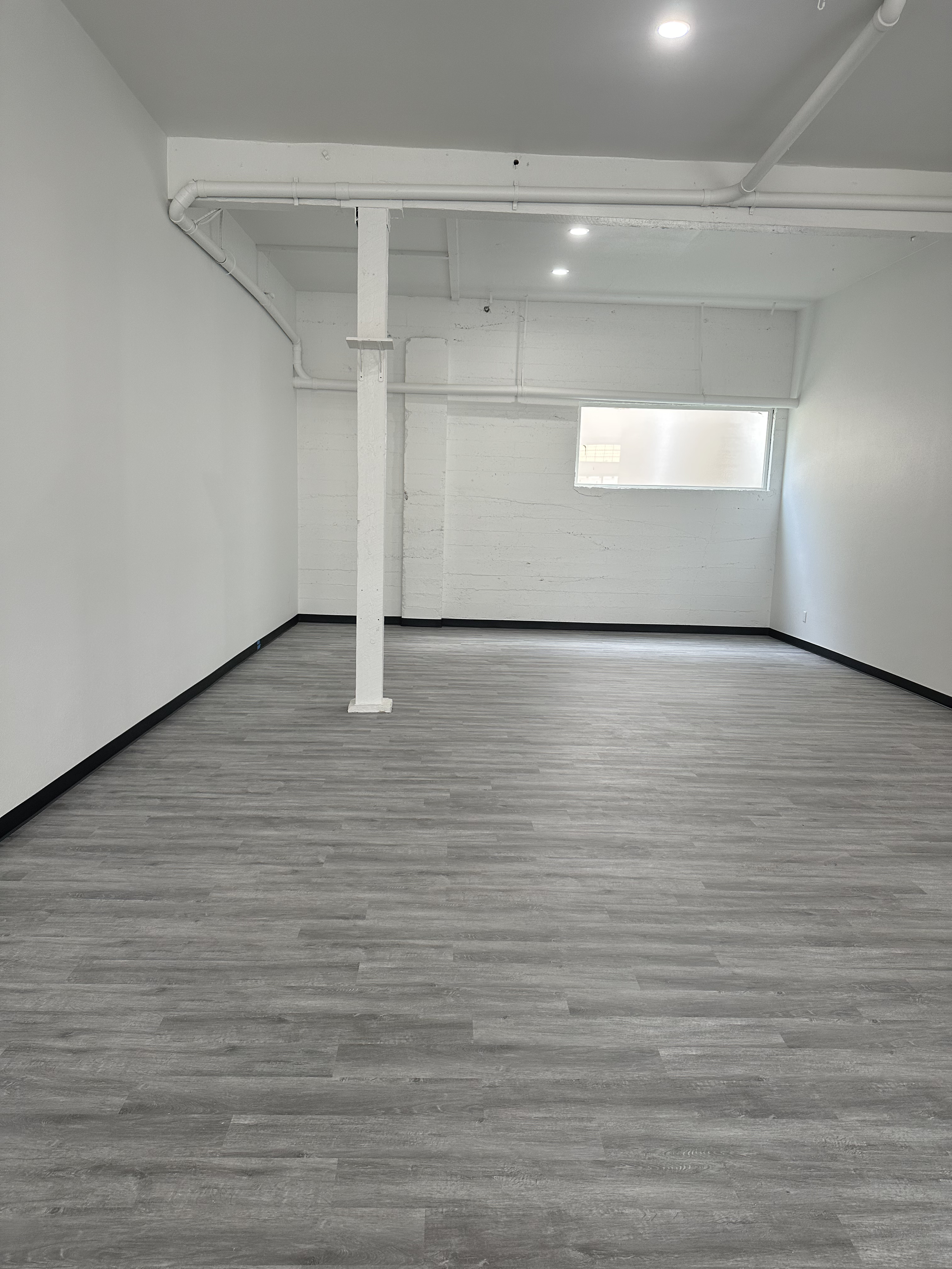 Empty room with gray wood-like flooring, white walls, black baseboards, a white painted exposed column, and ceiling pipes with recessed lighting, window on far wall.