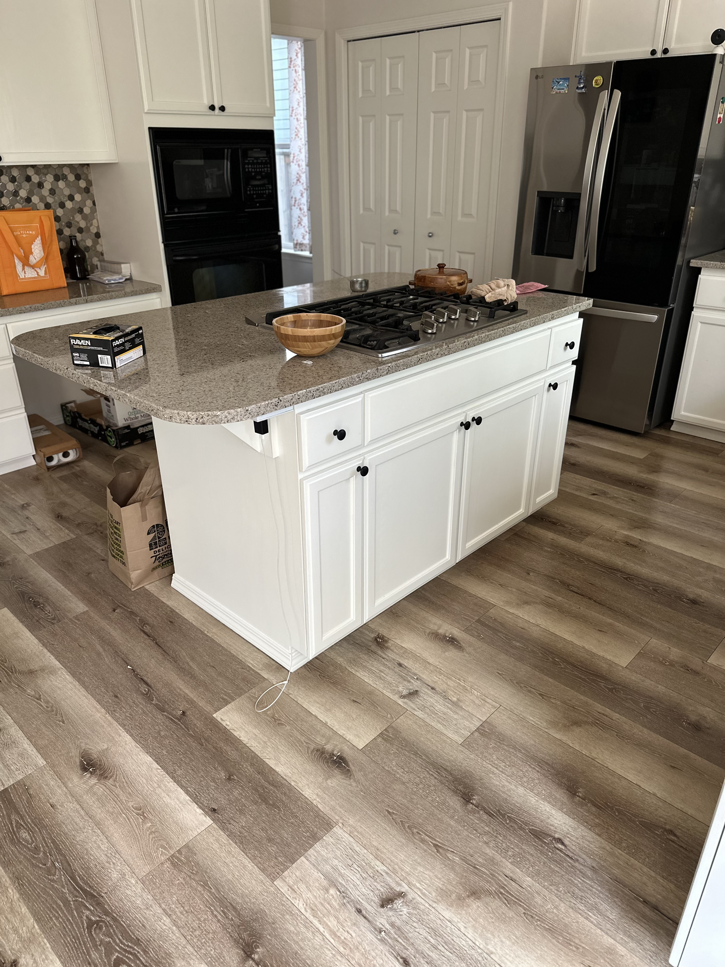 White kitchen island with a wooden bowl, some kitchen items, and a box on the countertop, with white cabinets, stainless steel refrigerator, and a gas stove in a modern kitchen.