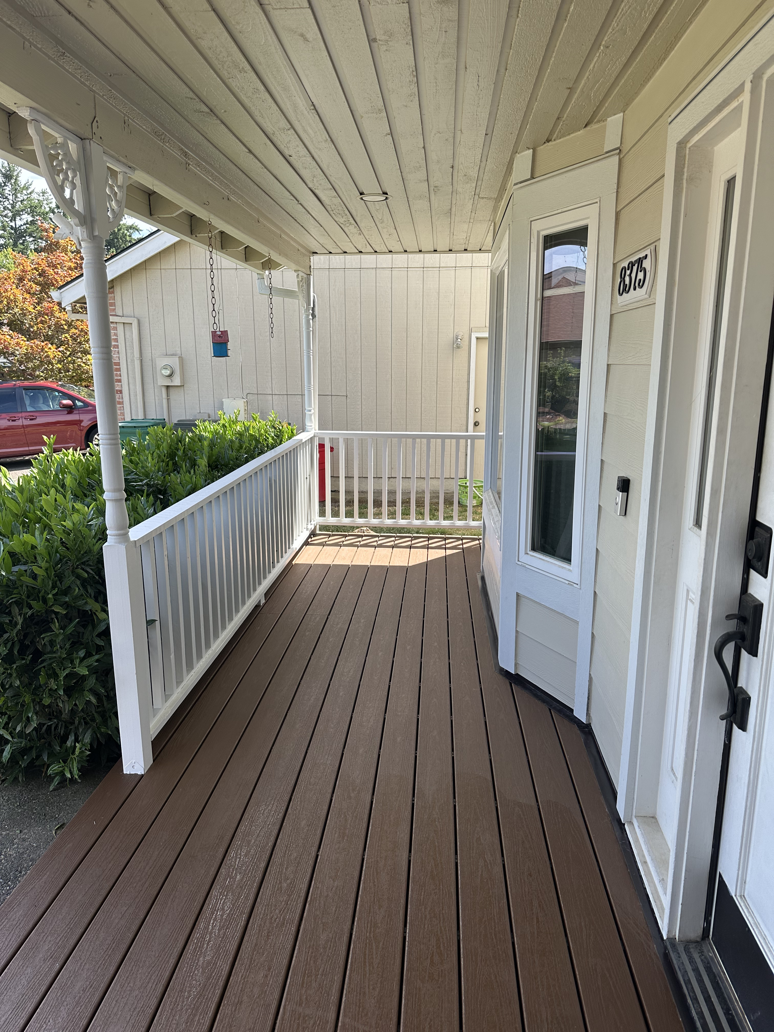 Front porch with brown wooden decking, white railing, and decorative white columns. A bush on the left side, a beige house with a window, and a white door with house number 8375 visible. A red car and trees can be seen in the background.