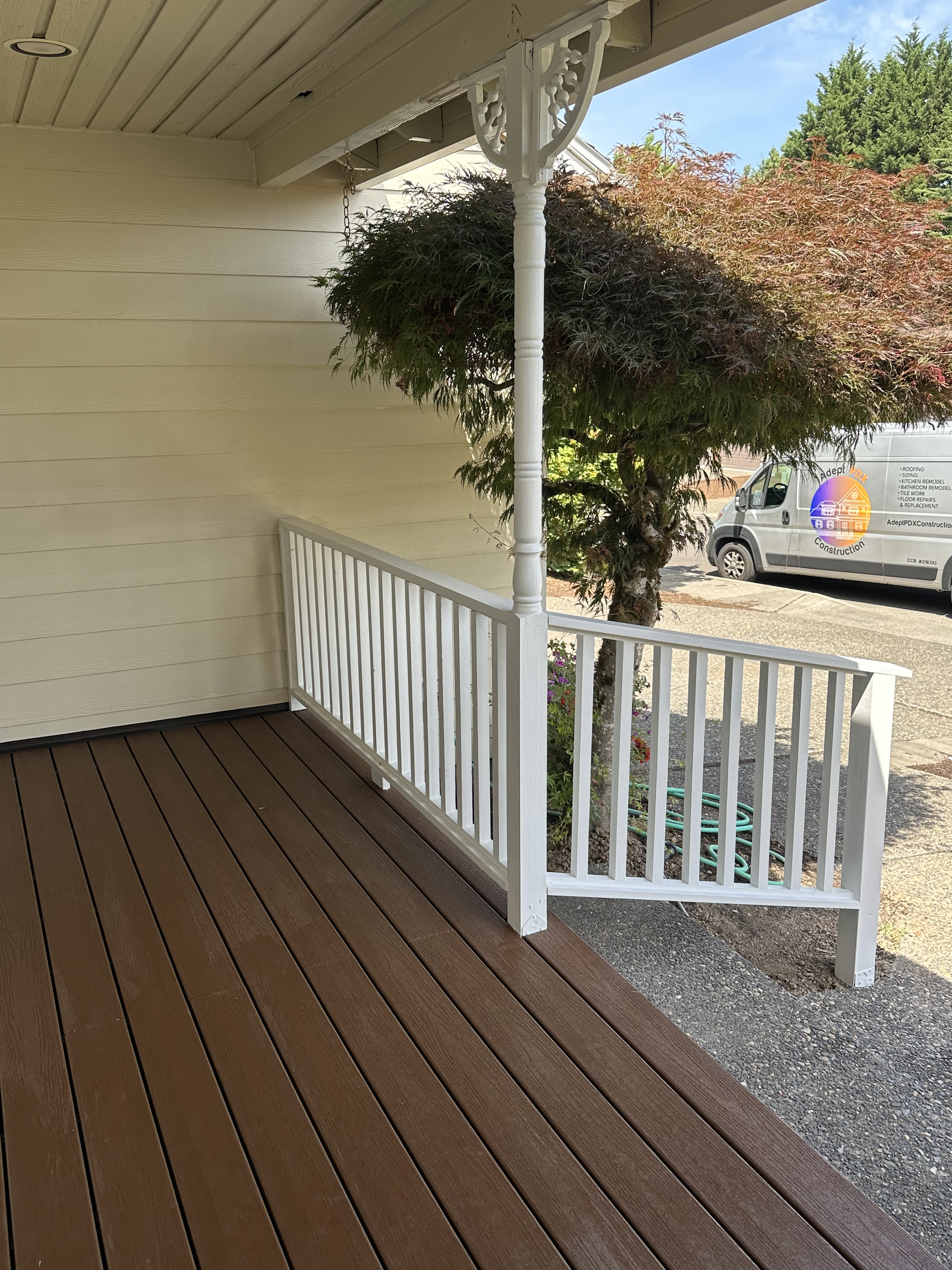 A porch with brown wooden deck flooring, white railing, an ornate white support post, a small garden with colorful flowers, a large leafy tree, a parked van on the street, and a sky with some clouds.