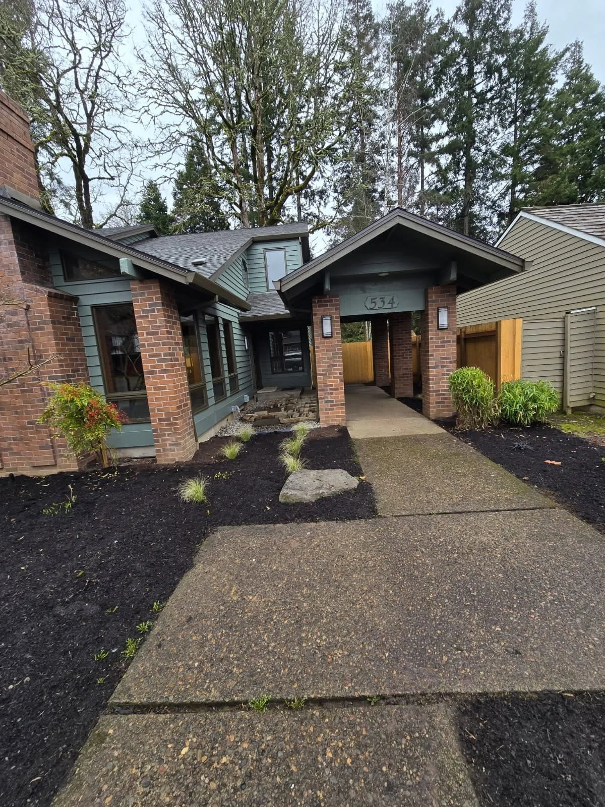 Front view of a modern house with brick and siding exterior, a paved walkway leading to the front door, and landscaped yard with small plants and mulch.