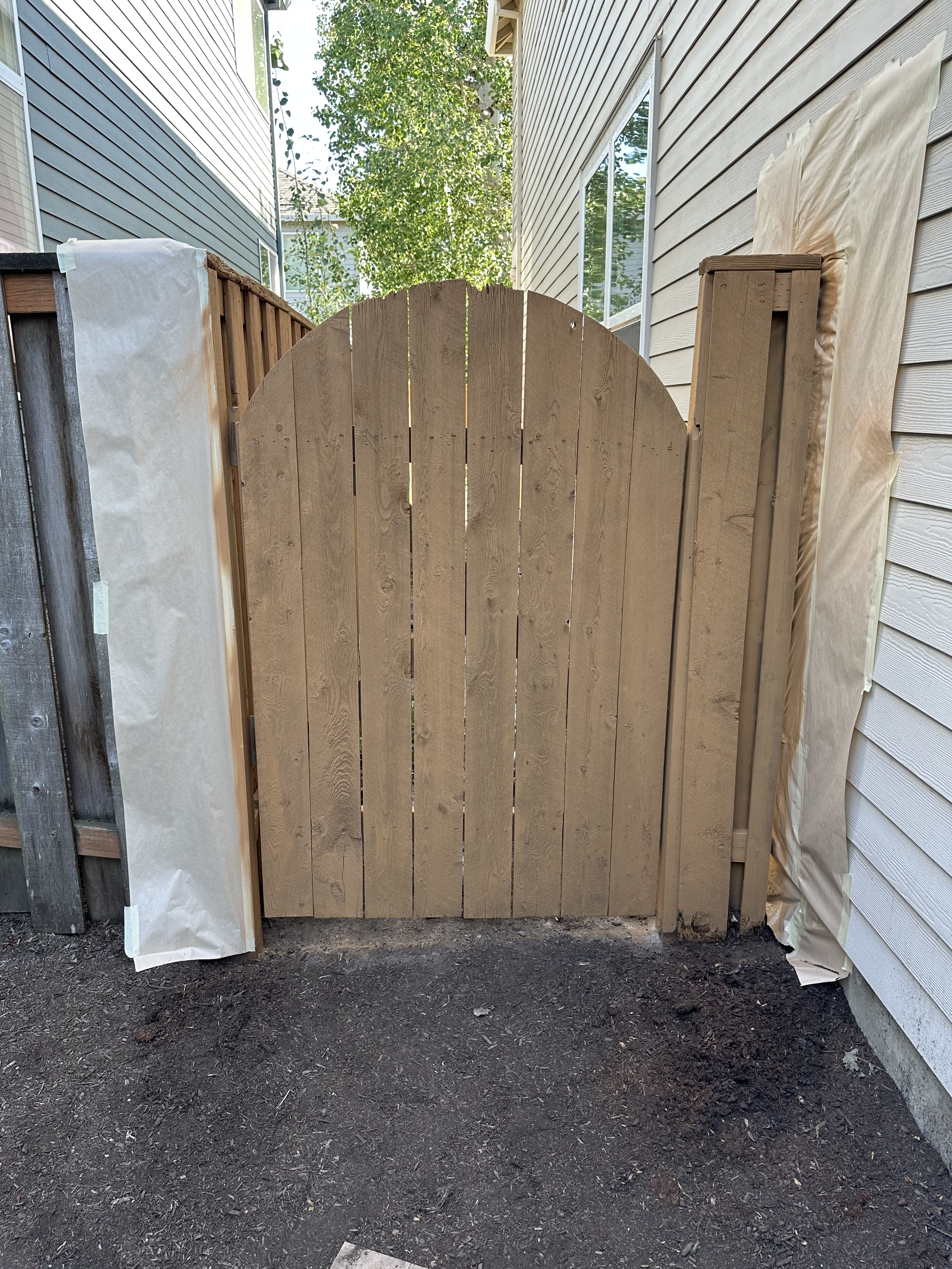 Wooden gate installed between two houses on dirt ground, with some protective wrapping on sides.