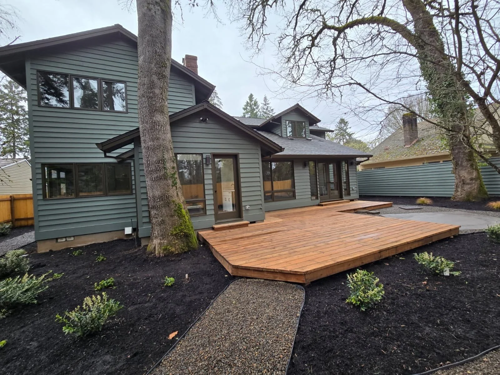 Backyard view of a gray house with a wooden deck surrounded by trees and new plants, overcast sky.