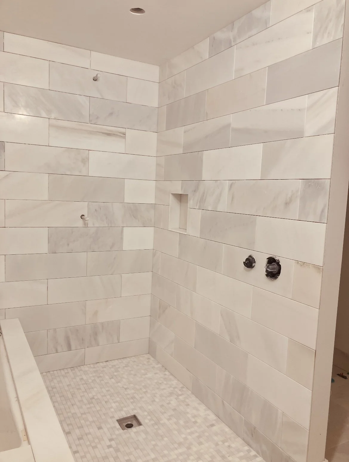 A newly constructed shower with white marble tiles on the walls, a small niche, and a tiled shower floor with a drain.