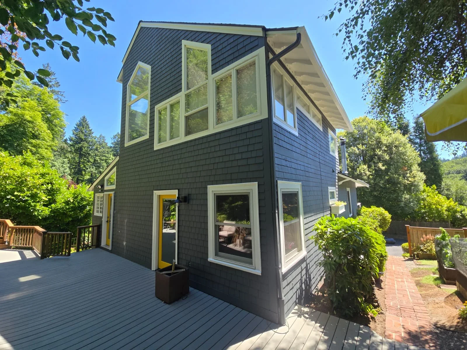 A multi-story house painted in dark gray with white window frames, surrounded by lush greenery and trees, and featuring a wooden deck with a yellow door and a potted plant outside.