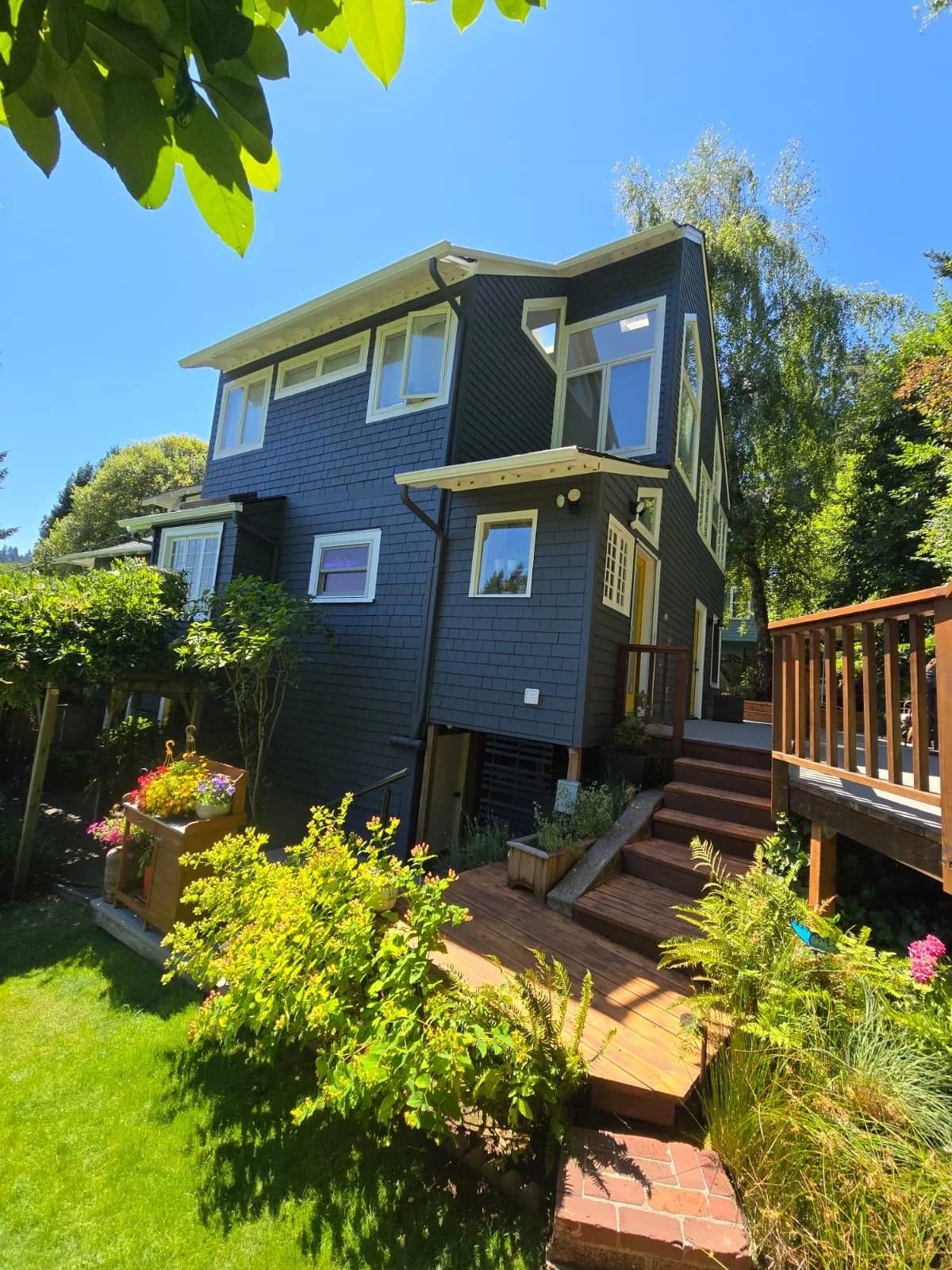 A modern, dark blue, three-story house with large windows, surrounded by greenery and a wooden deck.
